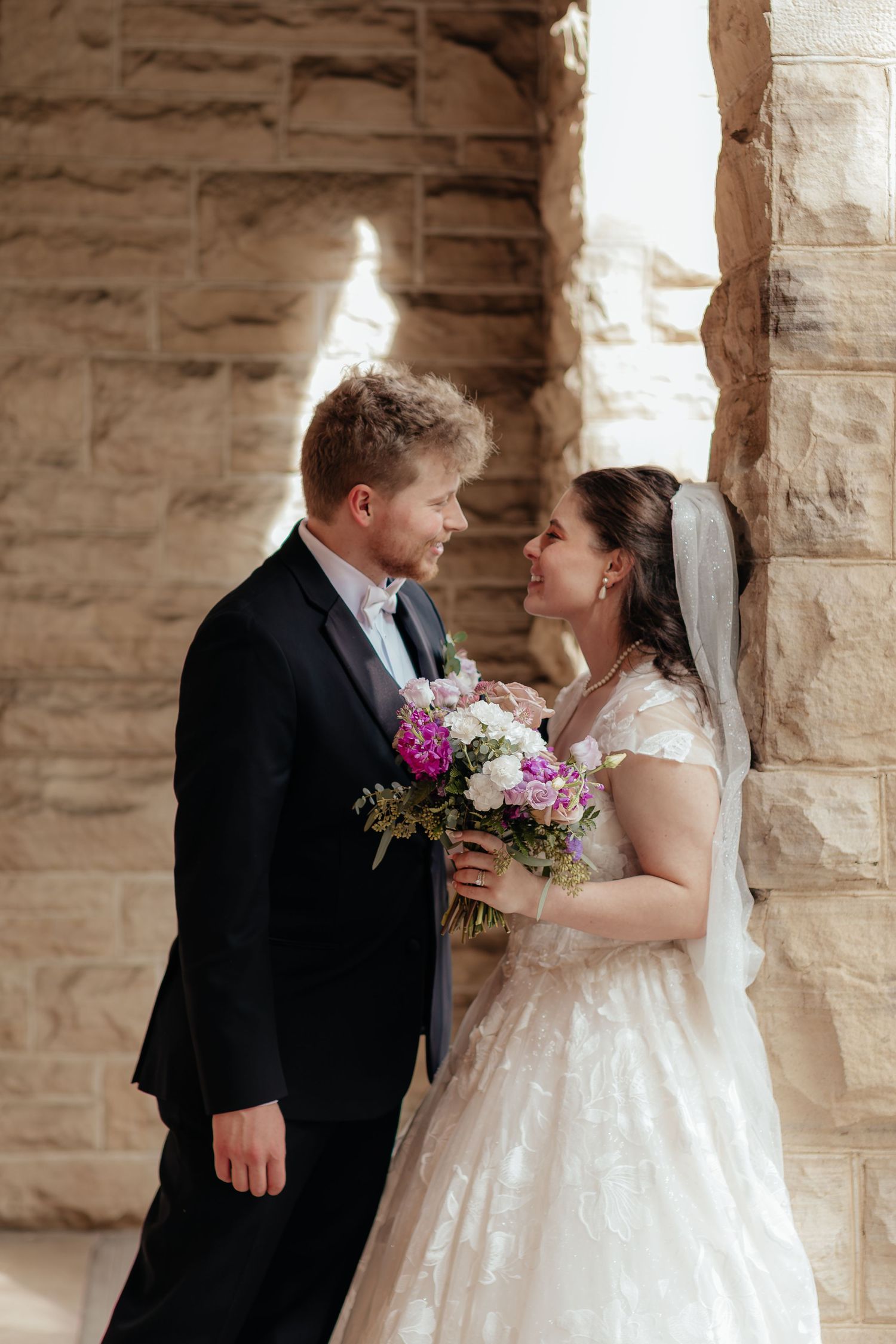Bride and groom share a romantic moment in front of a stone wall, bride holding a purple and white bouquet.