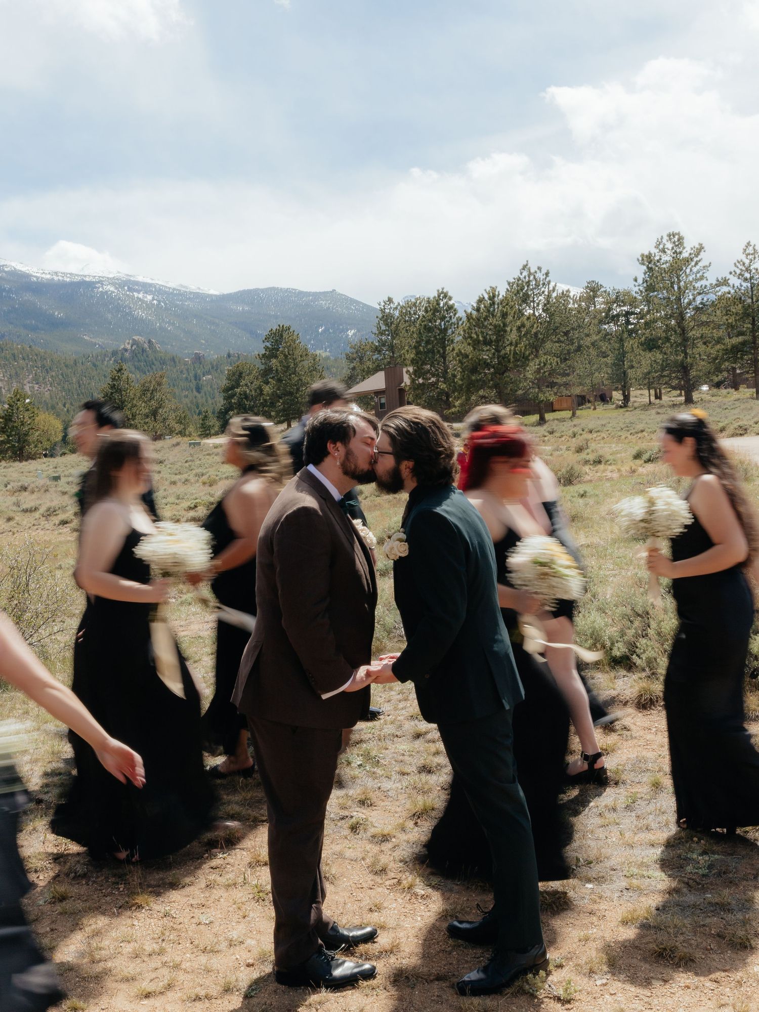 Two grooms share a kiss outdoors at a mountain wedding, surrounded by bridesmaids in black dresses.