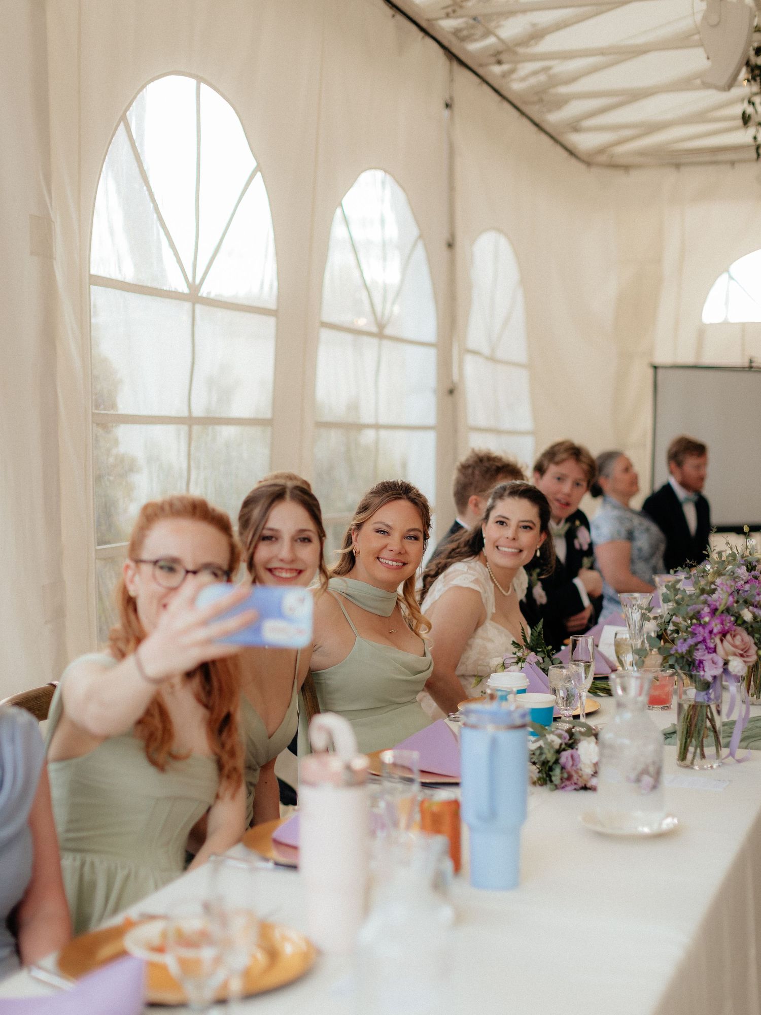Bridesmaids in sage green dresses taking a selfie at a wedding reception in a white marquee tent.