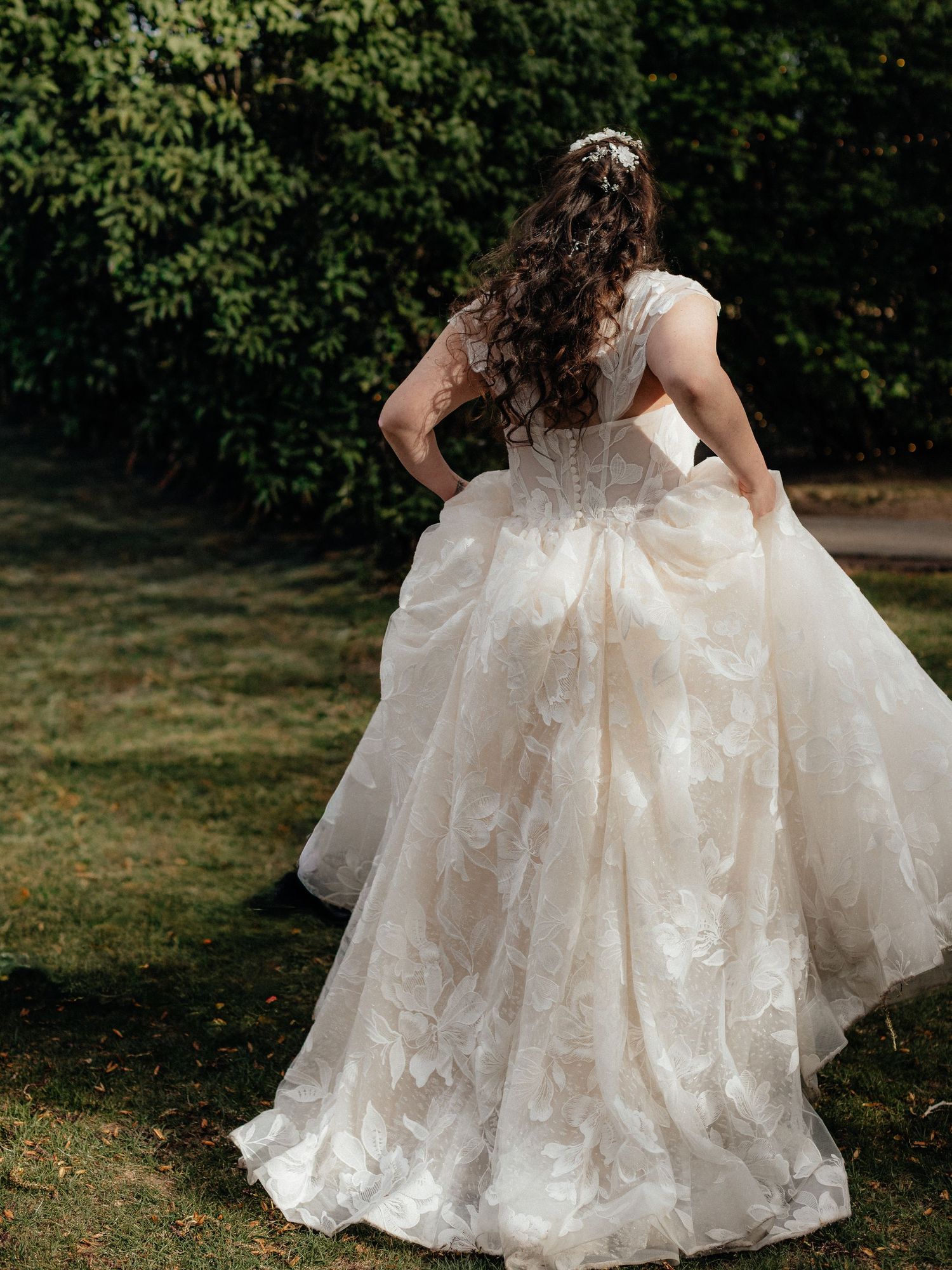 Bride in ornate lace ball gown with floral details walks through garden, showing stunning back view of dress.
