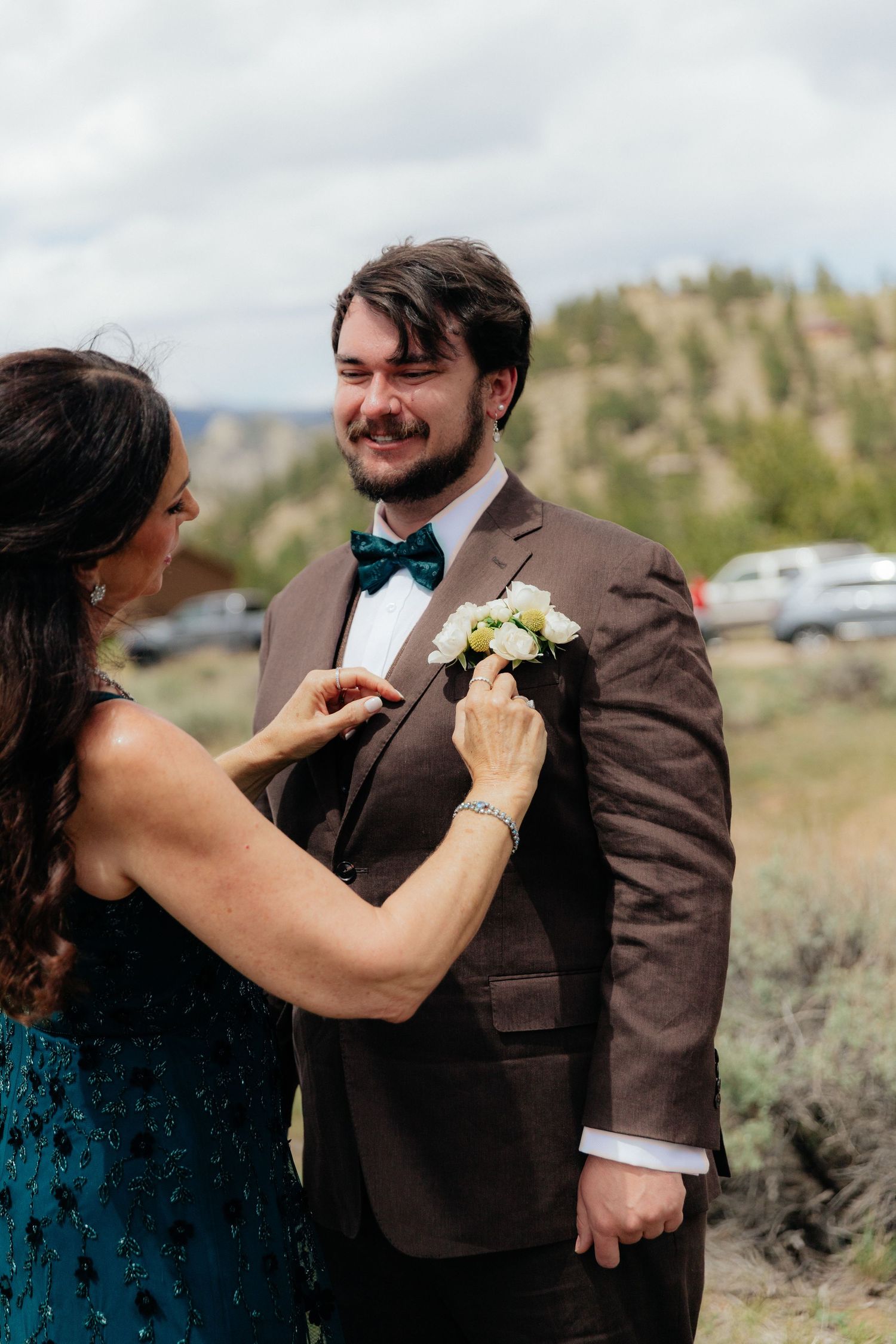 A woman in teal pins a white boutonniere on a smiling groom in a brown suit with teal bow tie outdoors.