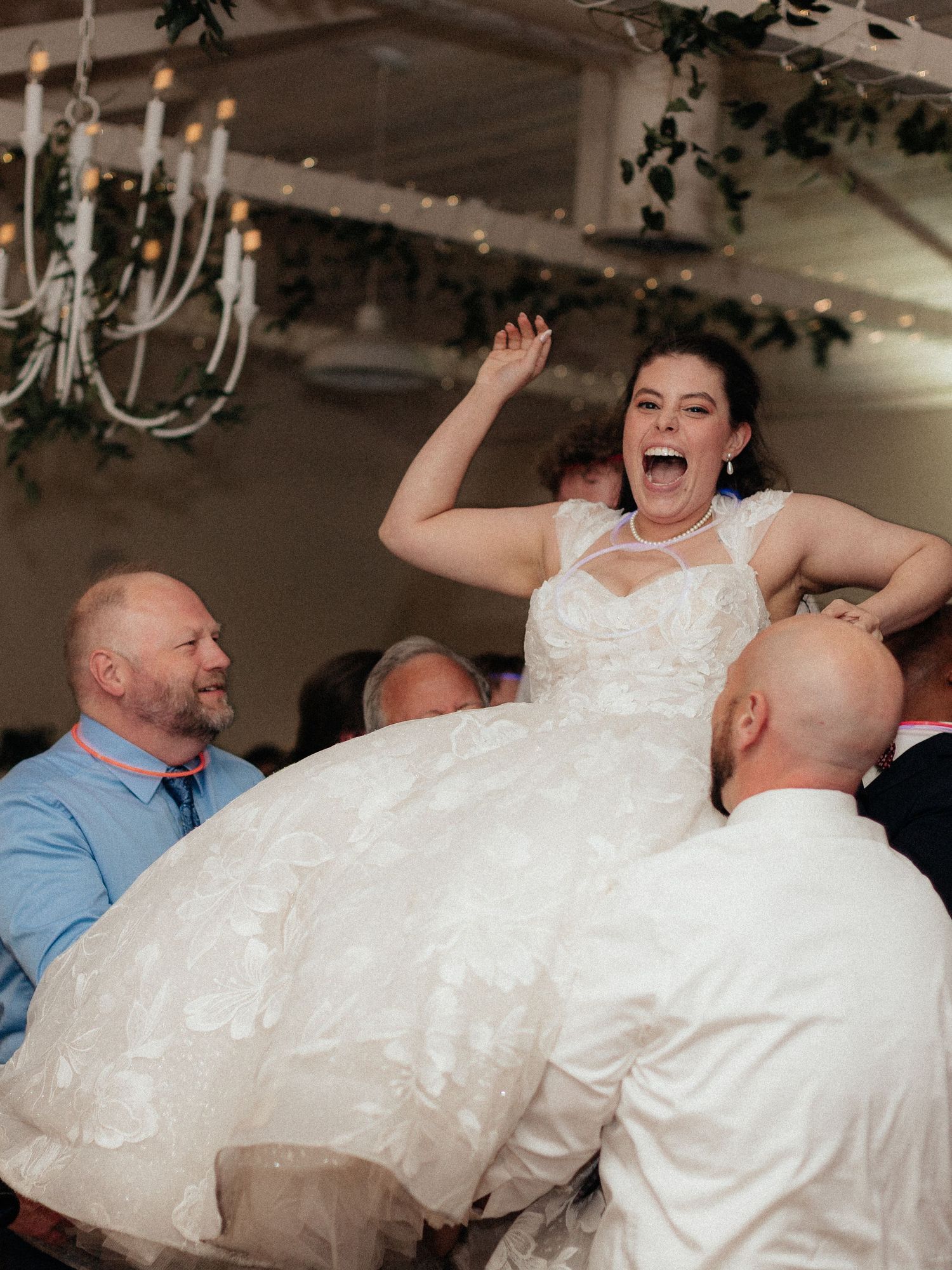 A joyful bride in a white lace gown is lifted by guests during a lively wedding reception.