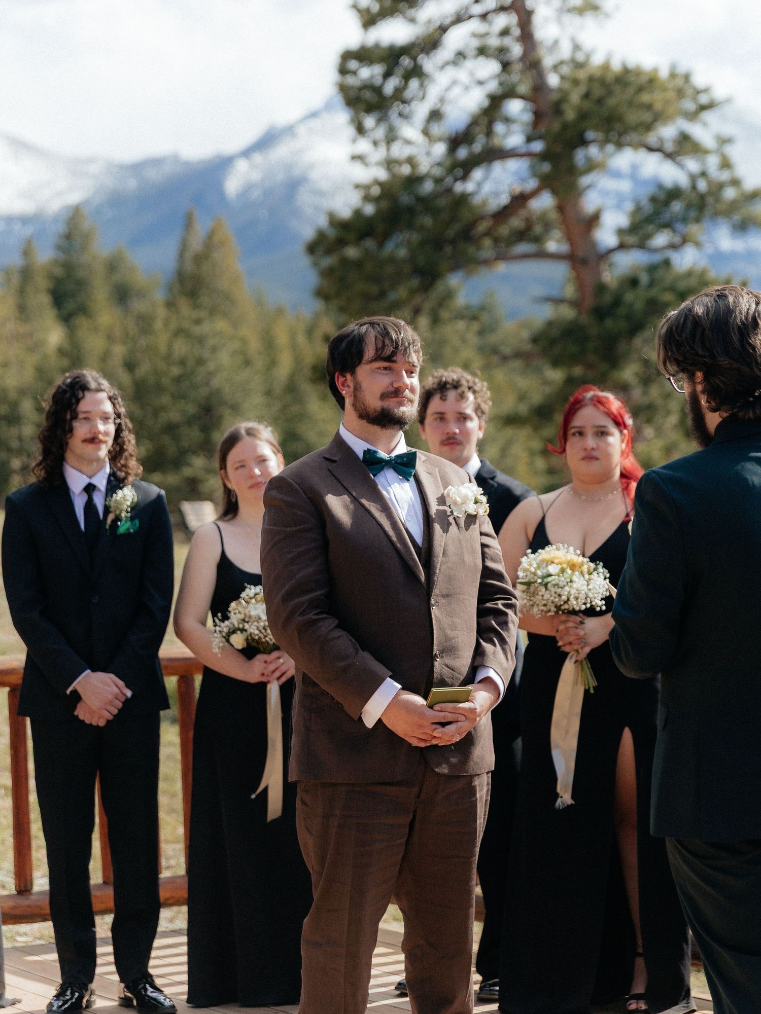 Groom in brown suit with bow tie stands at outdoor mountain wedding ceremony with wedding party in black attire.