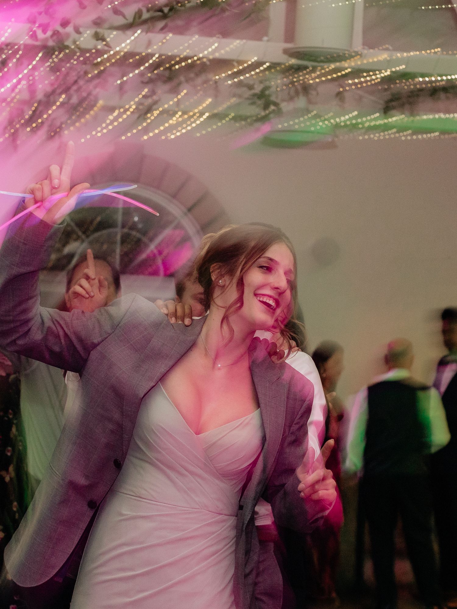 Bride in white dress dancing joyfully with guests under pink lights at a wedding reception celebration.