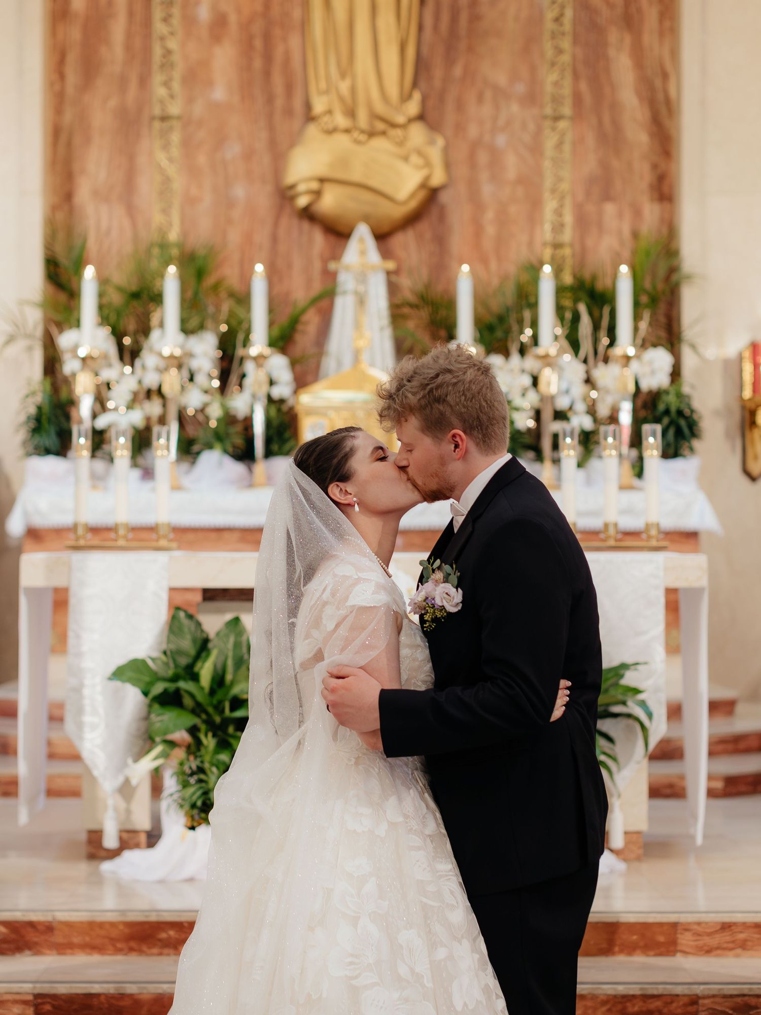 Bride and groom share first kiss at church altar, bride in white lace gown and veil, groom in black tuxedo.