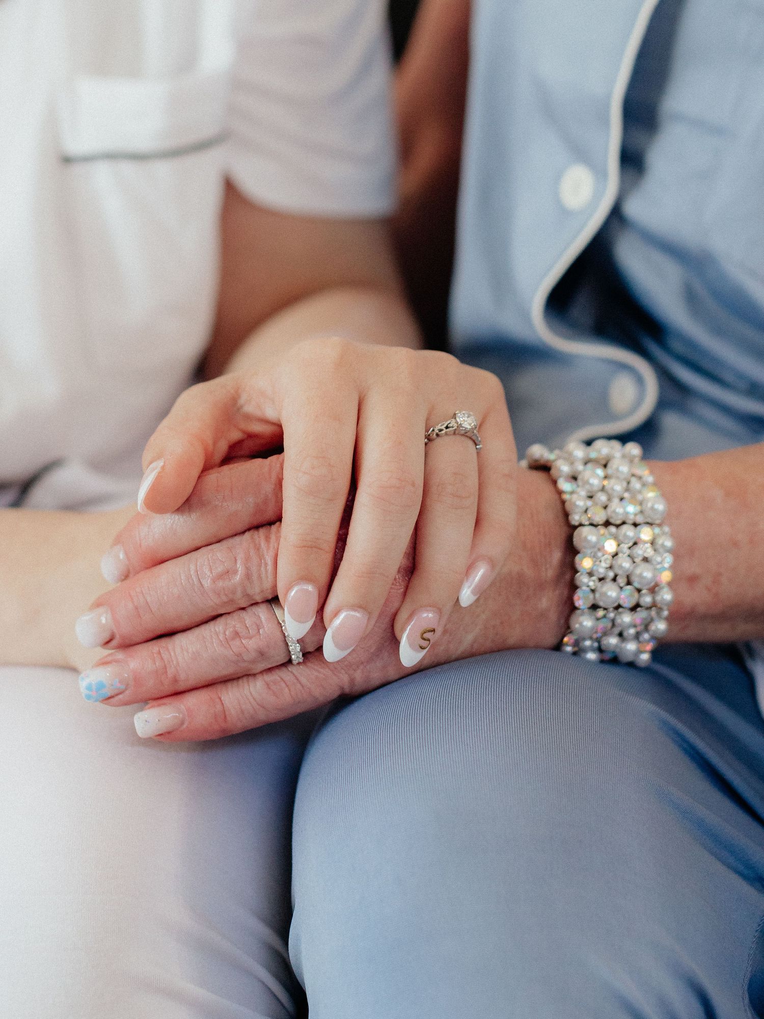 Close-up of two people holding hands, one wearing a diamond ring and pearl bracelet, with elegant white manicured nails.