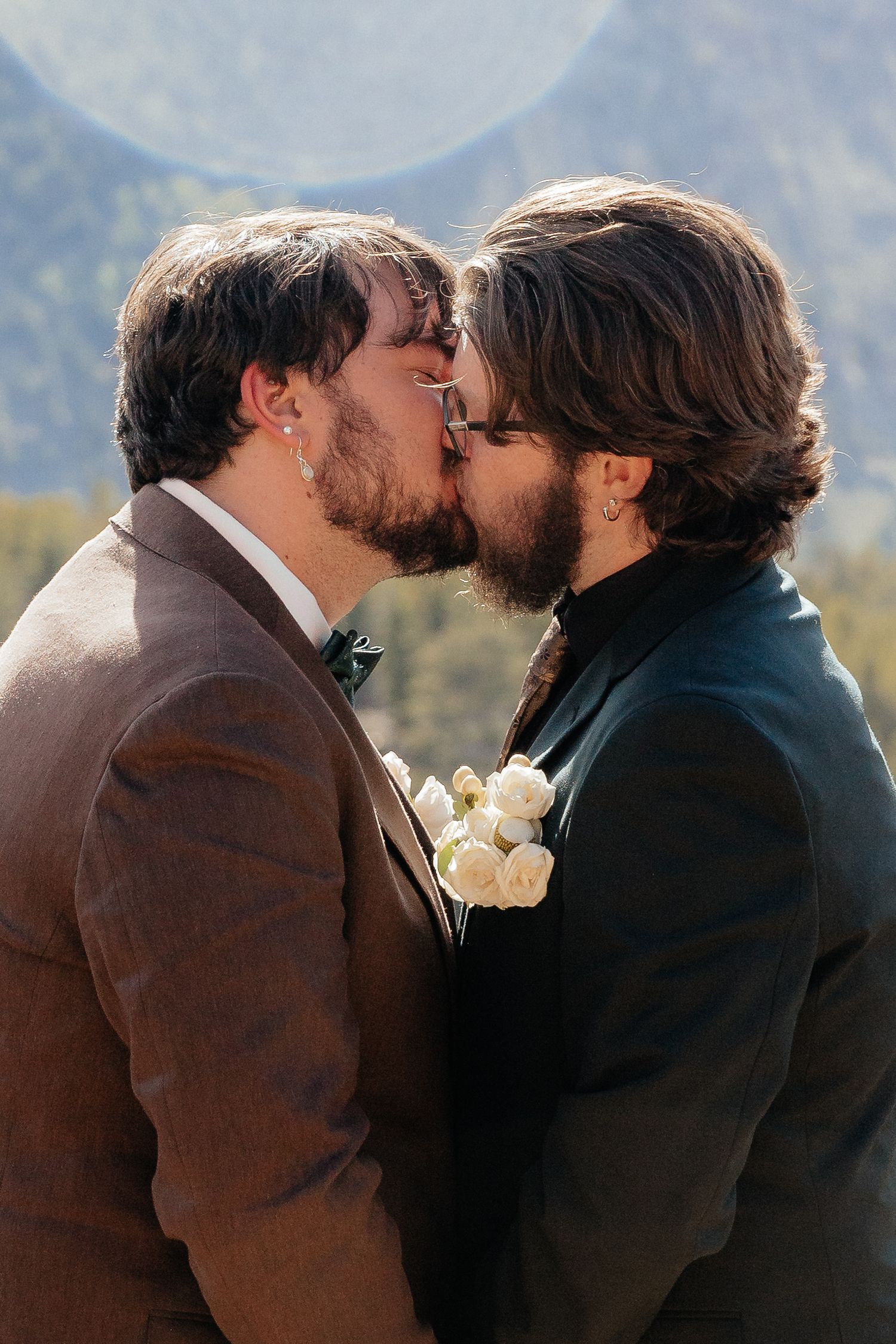 Two grooms sharing a first kiss at their wedding with mountain scenery in the background.