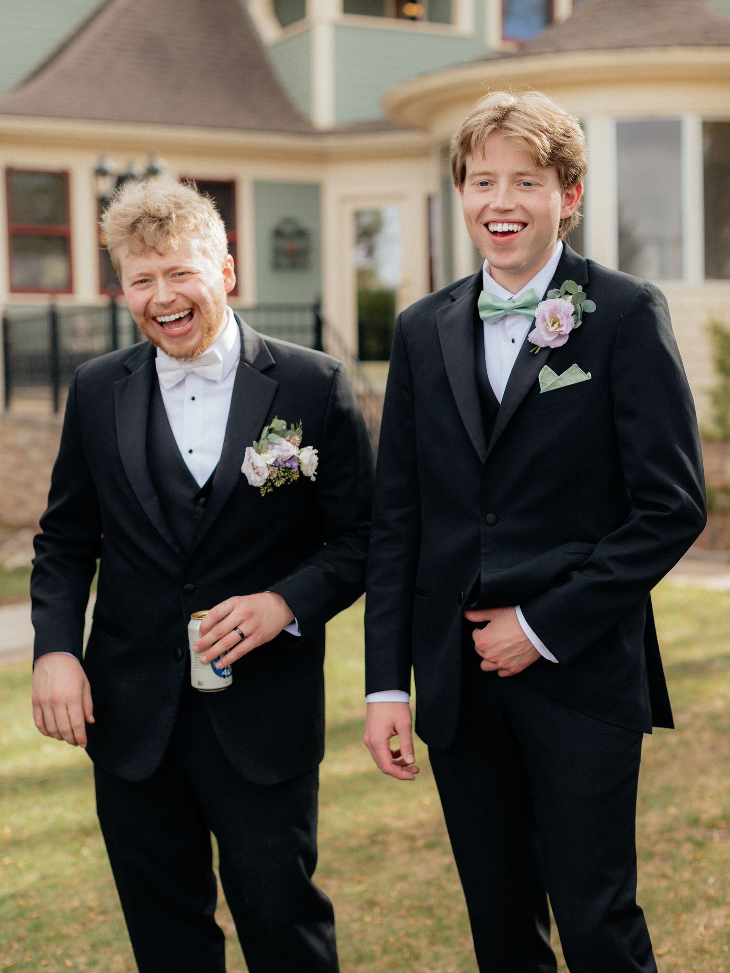 Two young men in black tuxedos with floral boutonnieres laugh joyfully outdoors at a formal event.