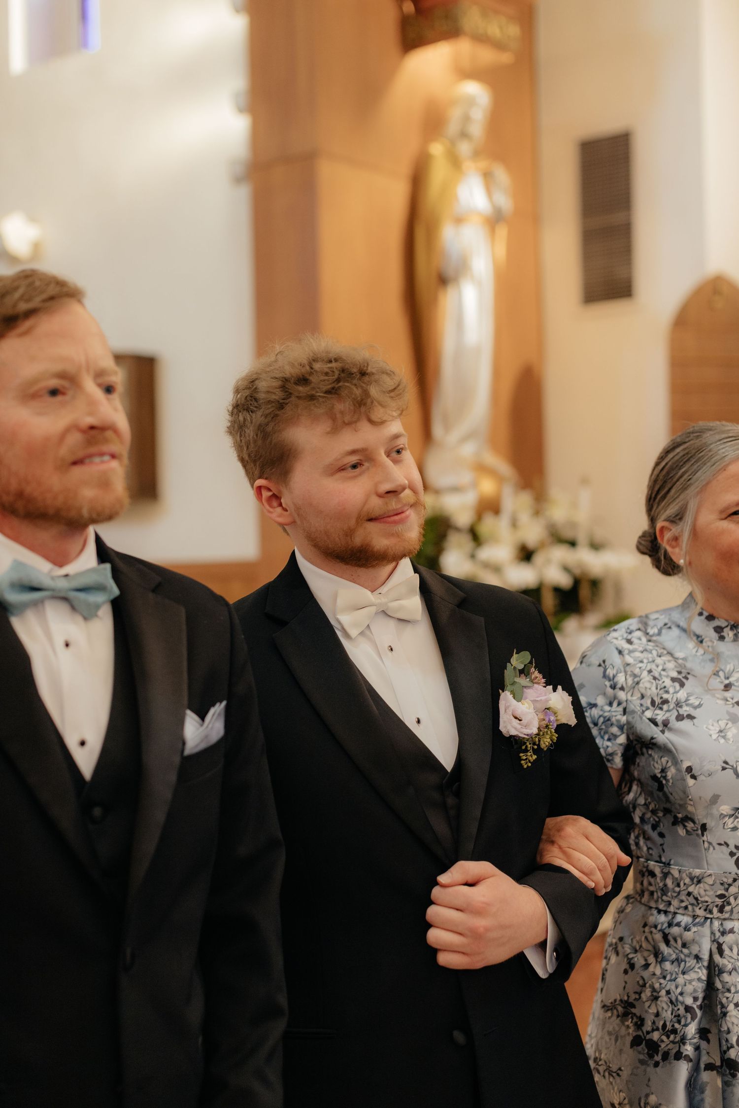 A groom in a black tuxedo with white bow tie stands at the altar flanked by family in a church.