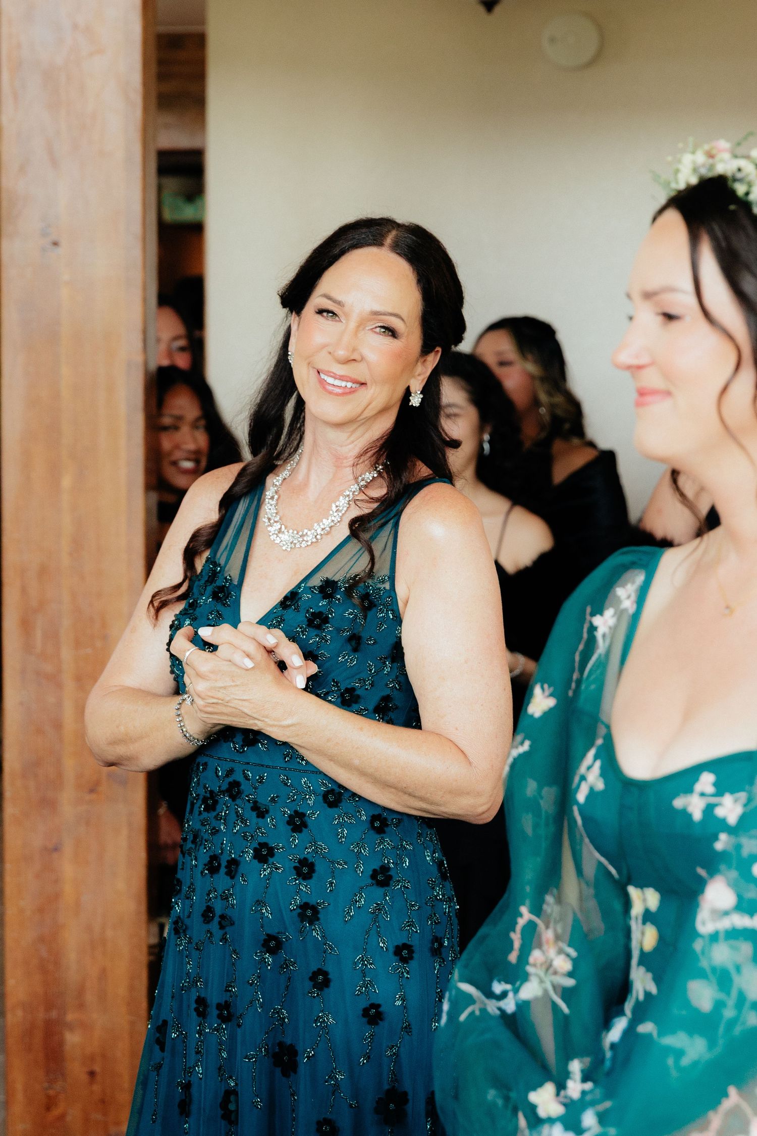 Elegant woman in teal beaded gown with diamond necklace smiling at a wedding celebration with guests in background.