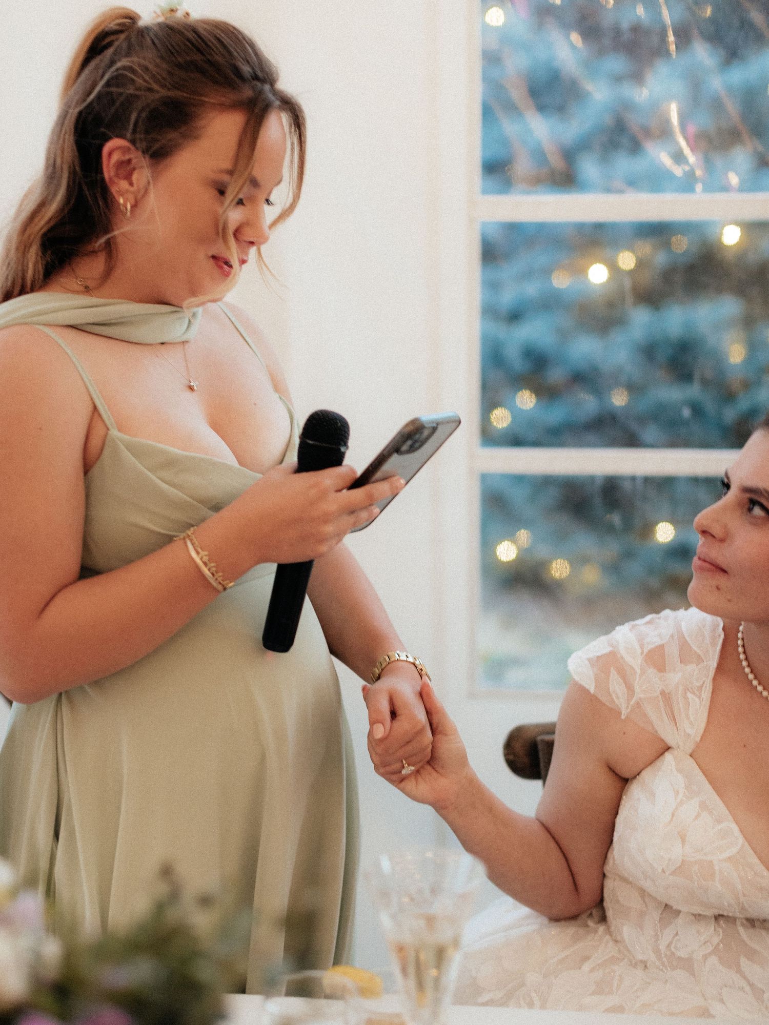 Bridesmaid in sage green dress holds microphone giving wedding speech while holding bride's hand at reception.