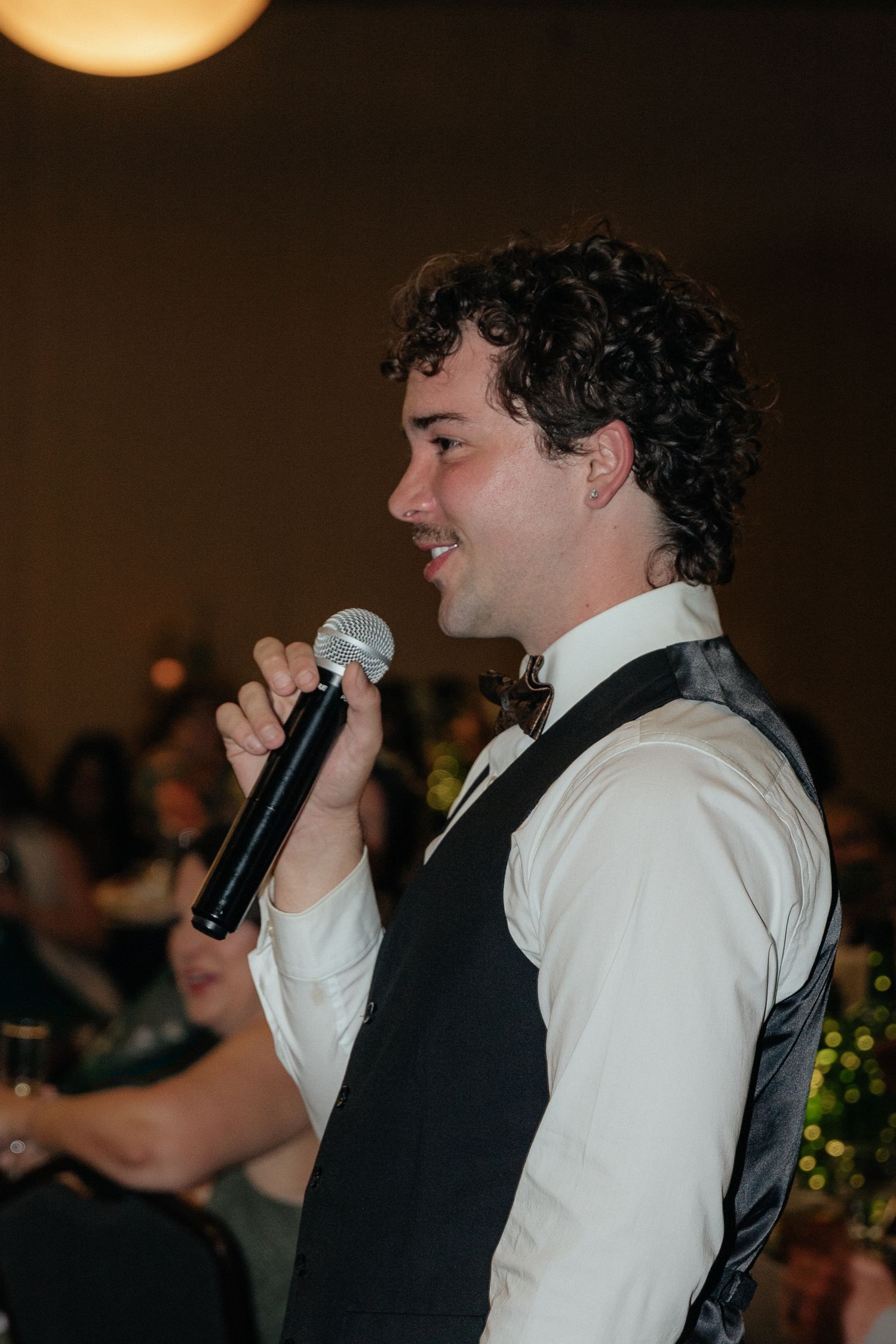Young man in vest and bow tie speaks into microphone at indoor wedding reception.