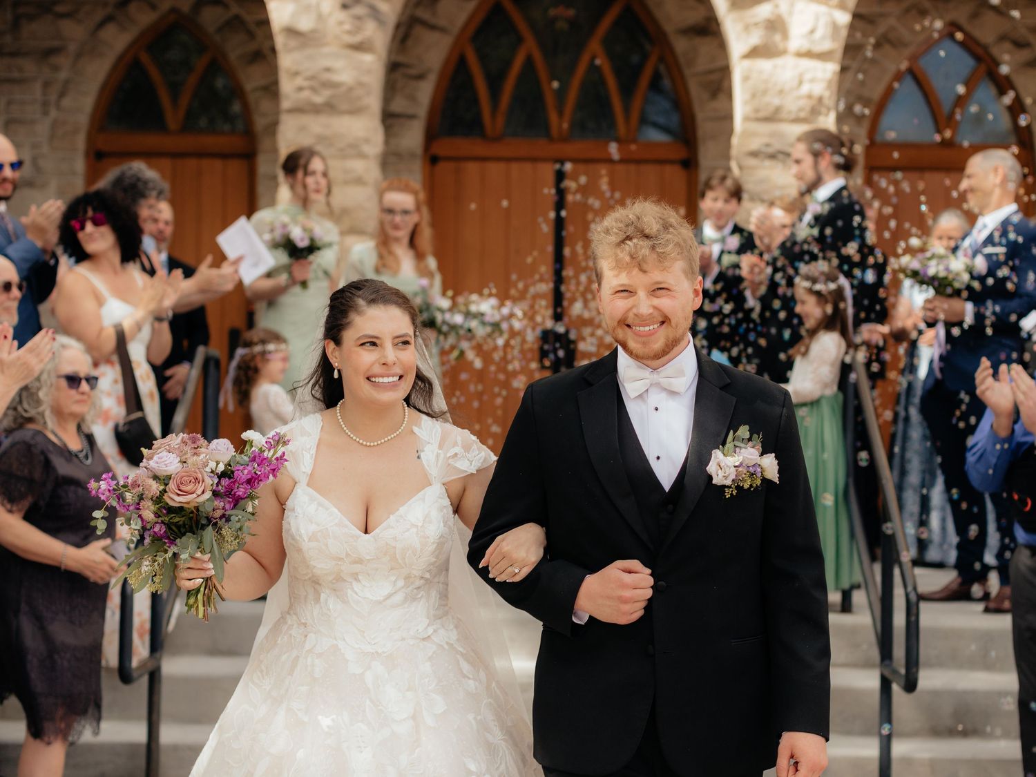 Newlyweds exit stone church amid confetti shower, bride in lace gown holding purple bouquet, groom in black tux.