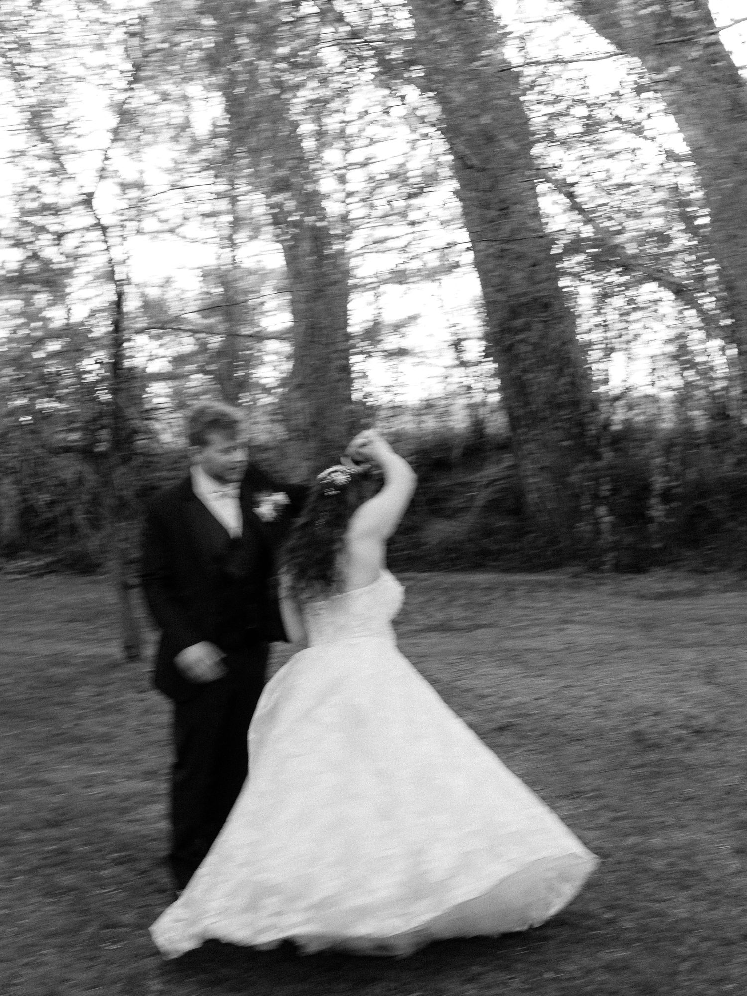 Black and white photo of a bride and groom dancing outdoors among tall trees on their wedding day.