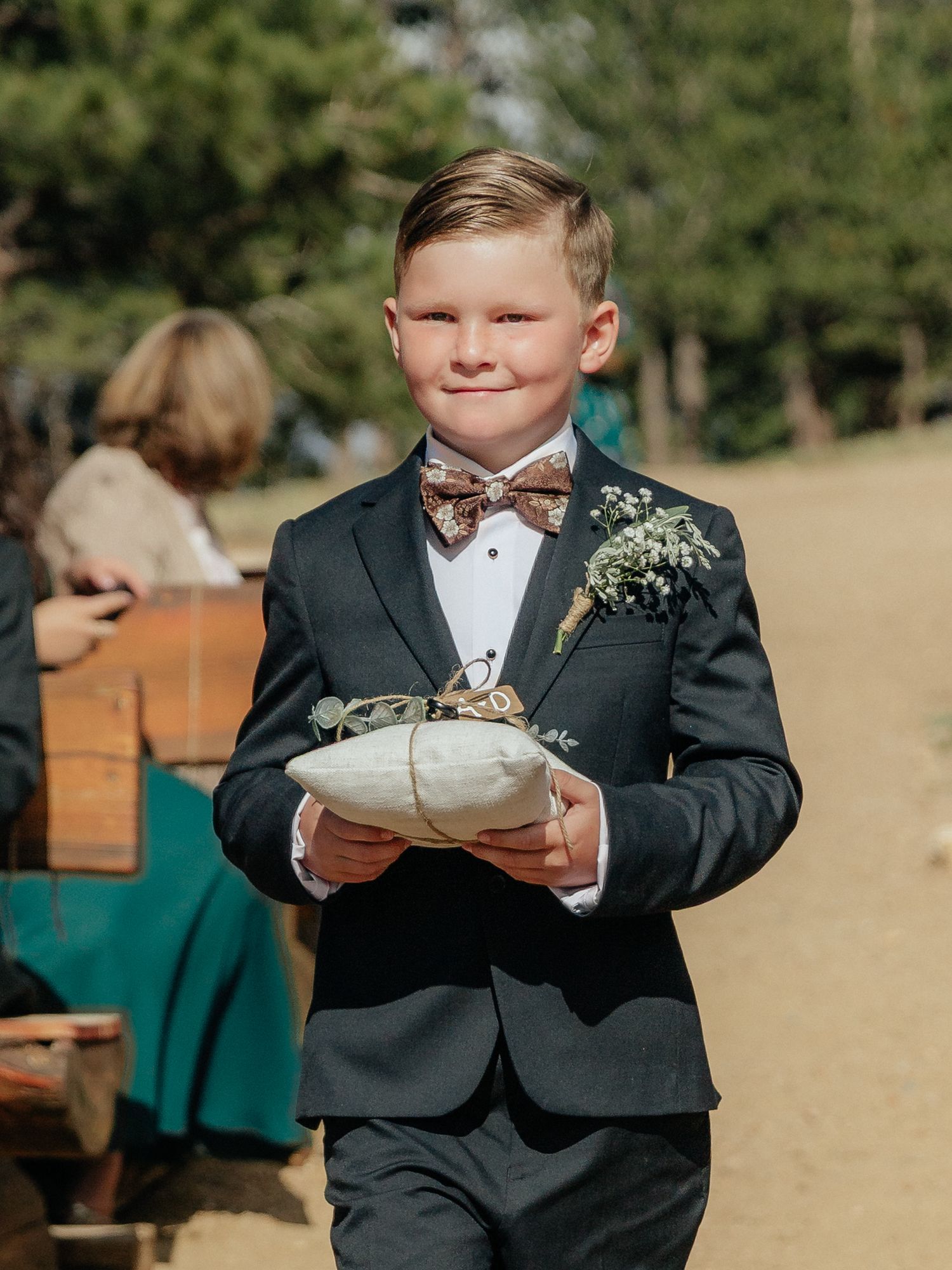 A young ring bearer in a dark suit and bow tie carries a pillow down the aisle at an outdoor wedding ceremony.