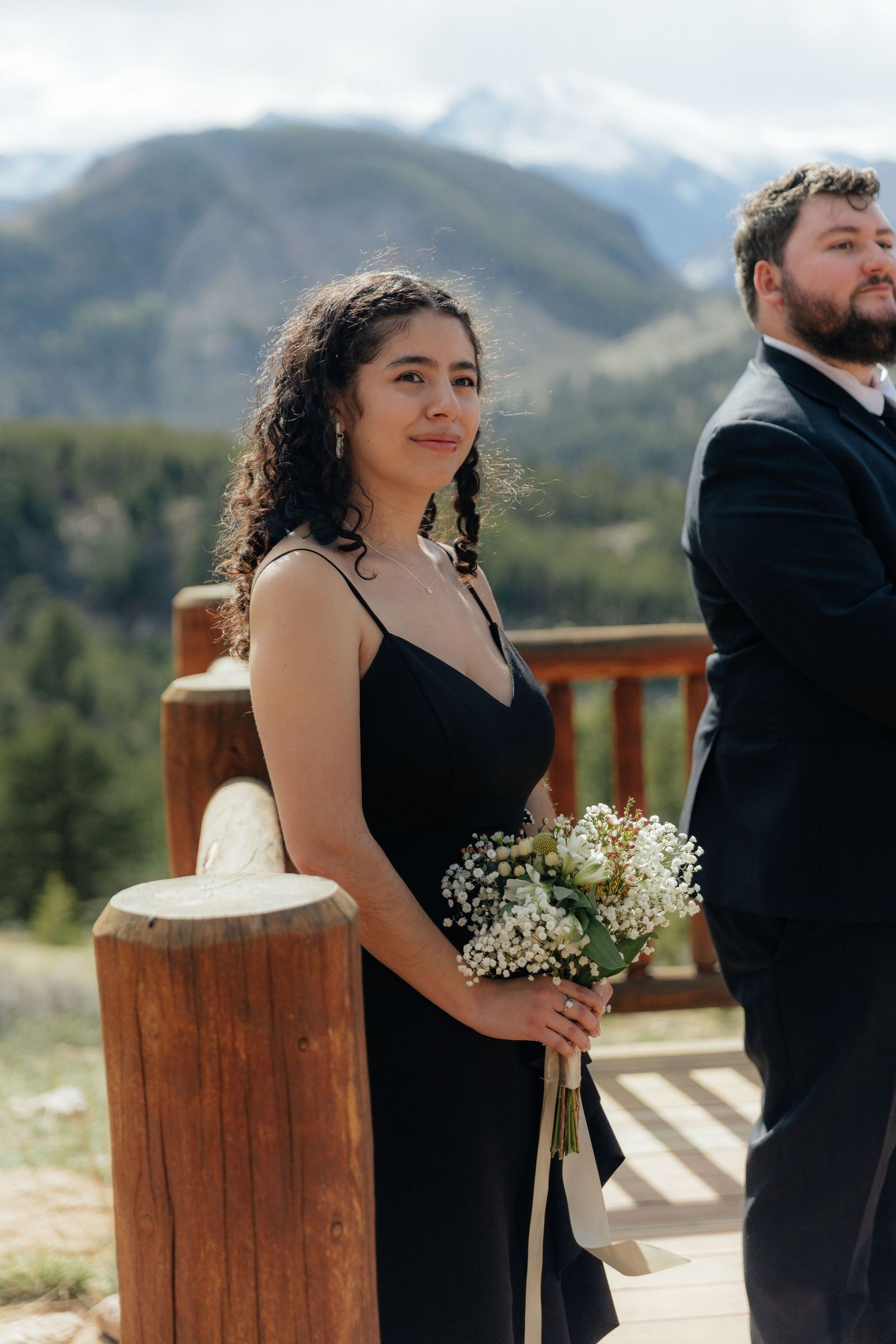 Bridesmaid in black dress holding white bouquet at mountain wedding ceremony on wooden deck.