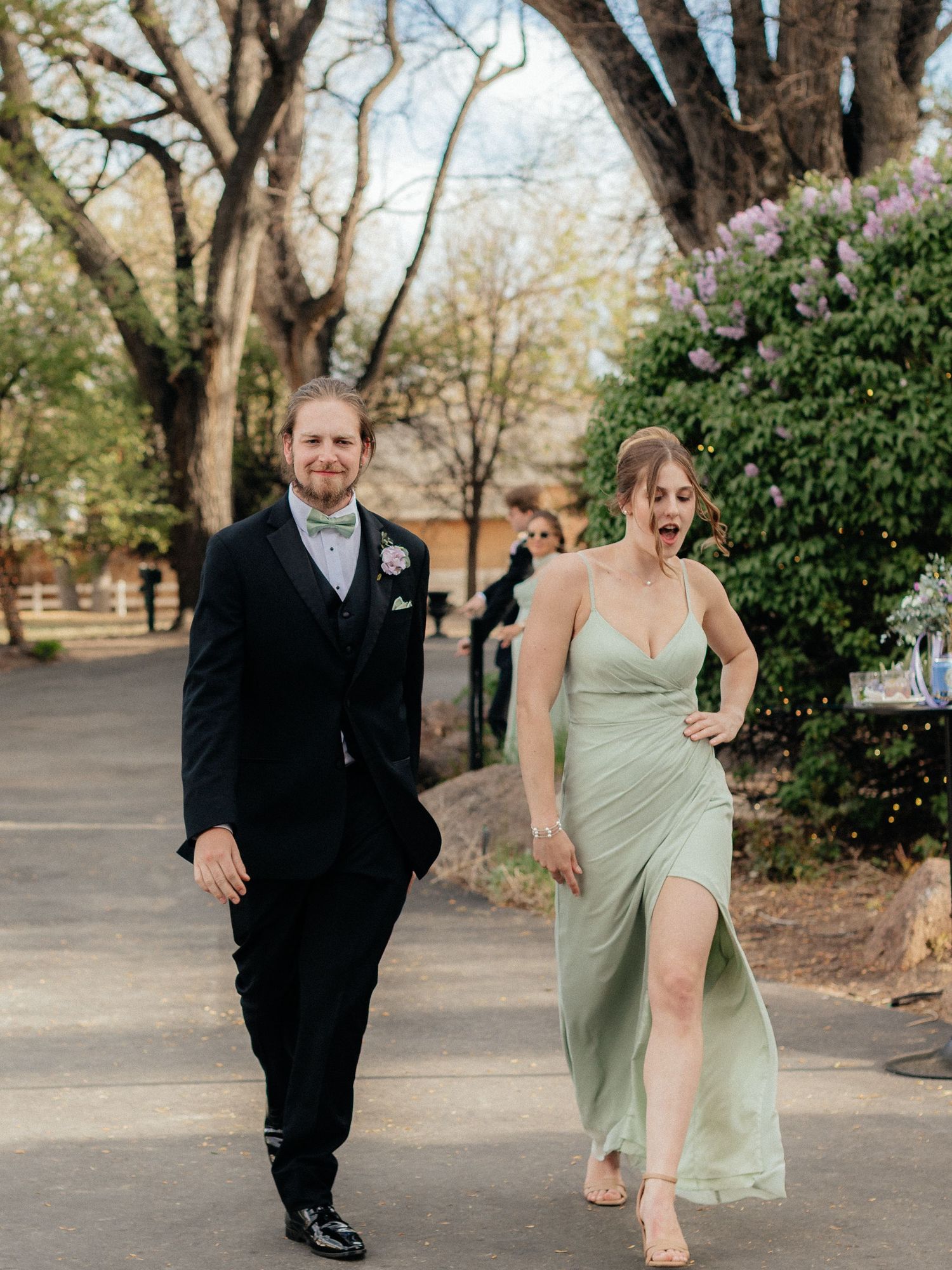 Smiling couple in wedding attire walking outdoors, man in black suit, woman in sage green slit gown.