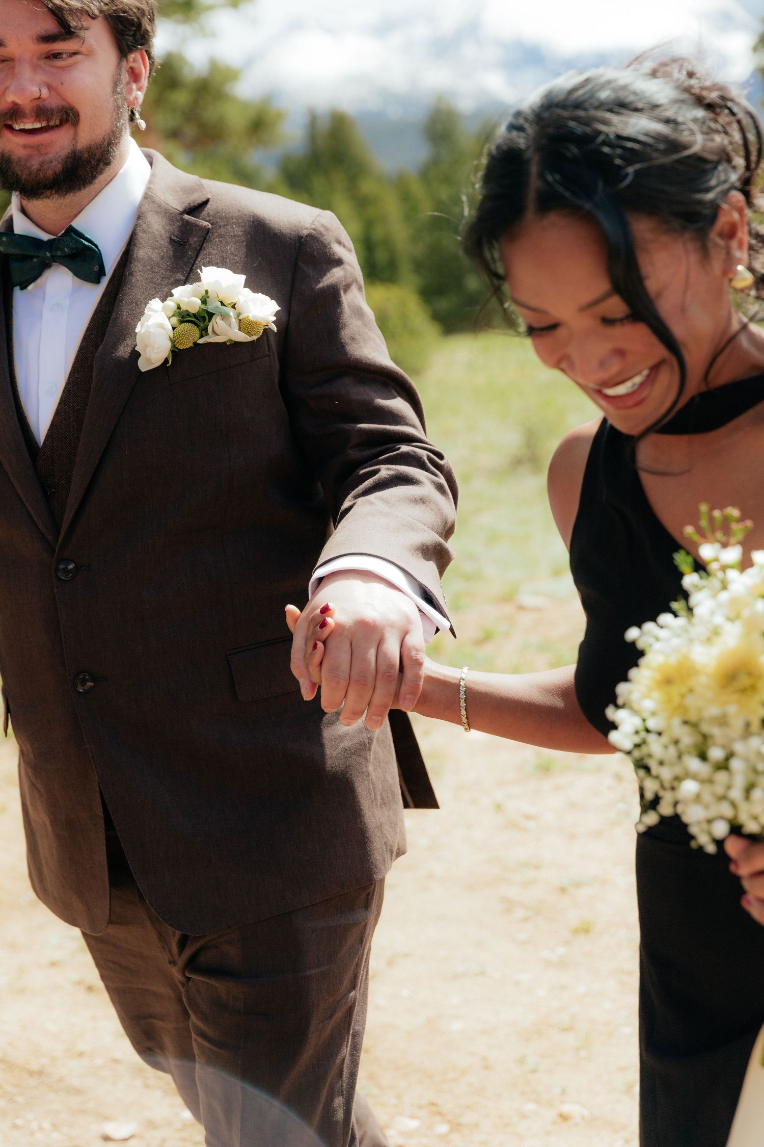 Smiling bride in black dress holds groom's hand outdoors, groom wearing brown suit with green bow tie and white boutonniere.