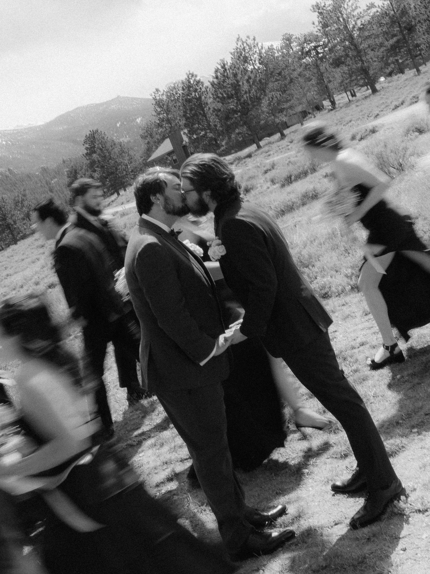 Two grooms share a kiss outdoors at a rustic mountain wedding in a black and white photo.