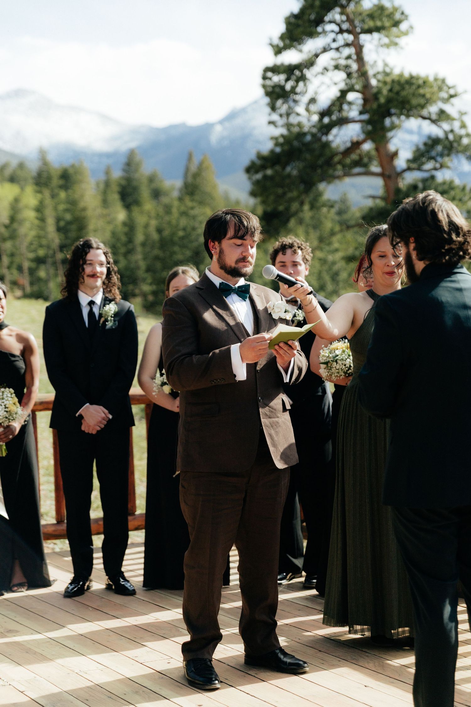 Groom in brown suit reading vows at mountain wedding ceremony on scenic outdoor deck.