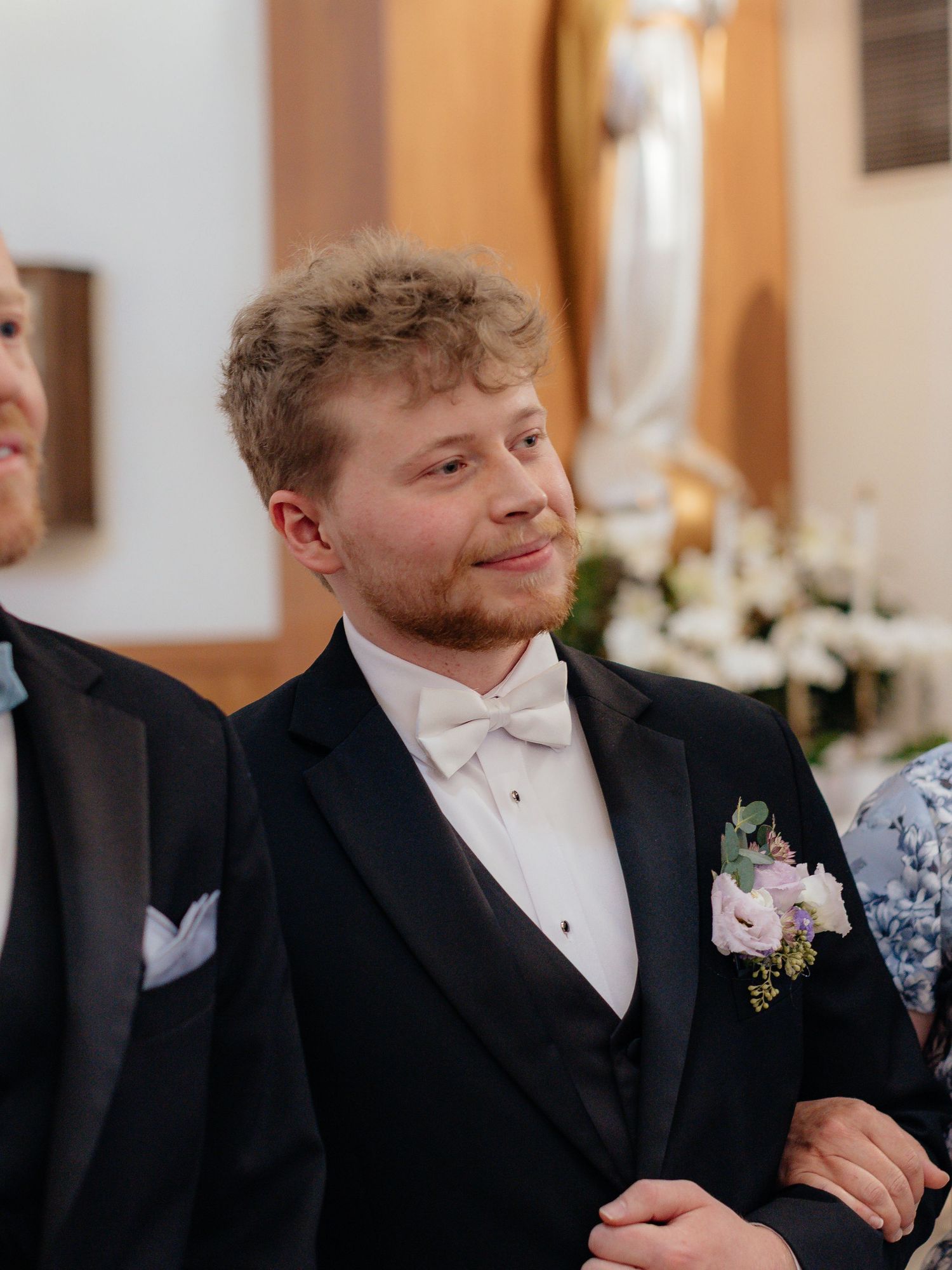 Young groom in black tuxedo with white bow tie and floral boutonniere smiling at a church wedding ceremony.