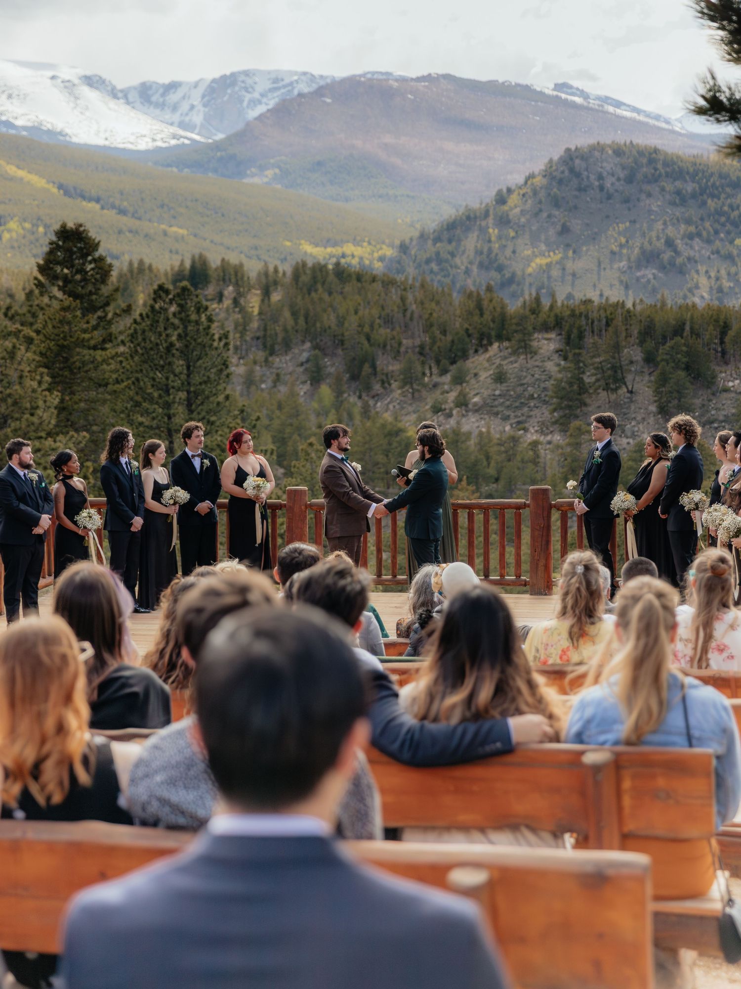 Outdoor mountain wedding ceremony on a wooden deck with bridal party in black attire and snow-capped peaks backdrop.