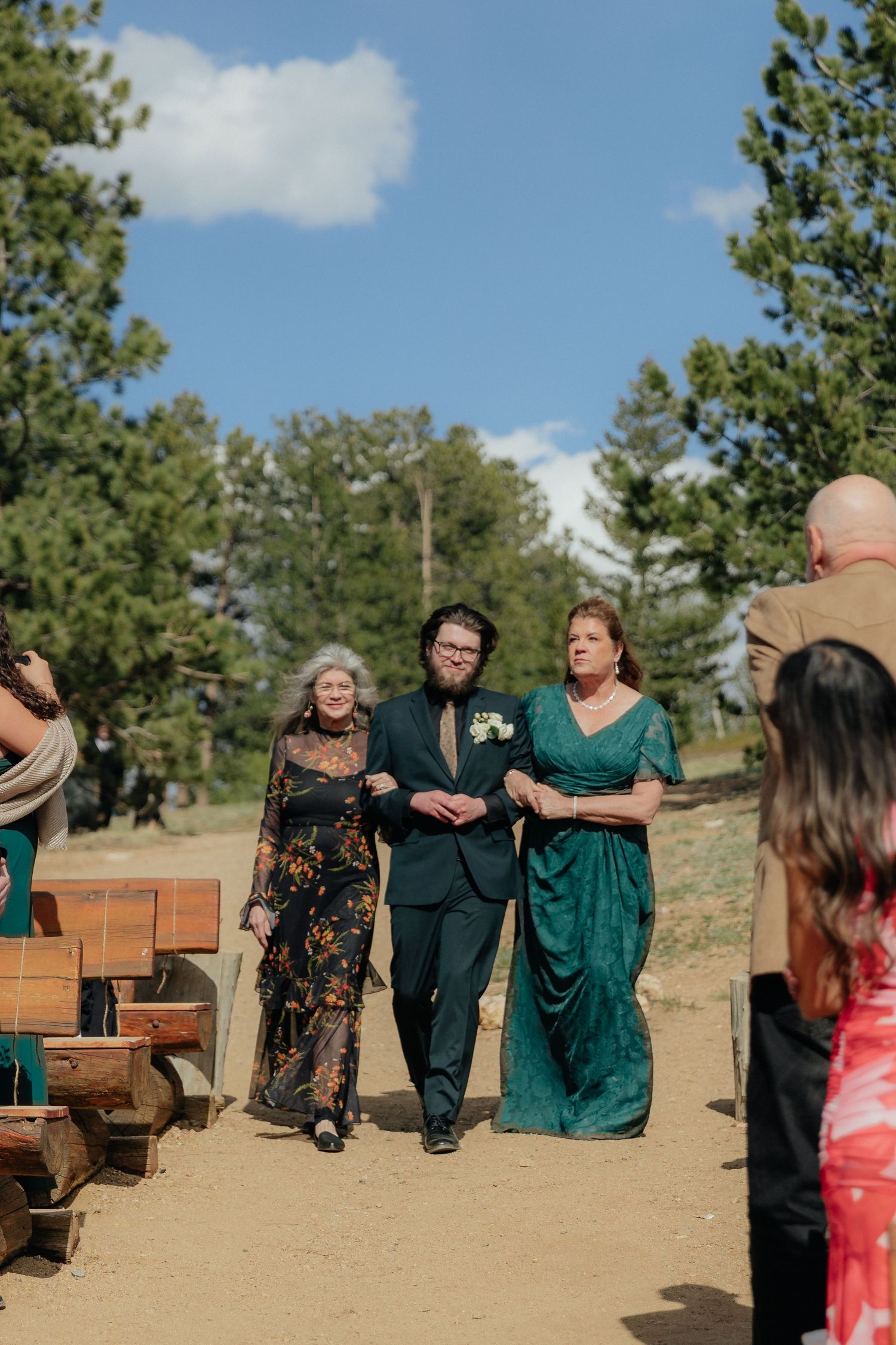 Groom walks down outdoor wedding aisle escorted by two women in green dresses among pine trees.