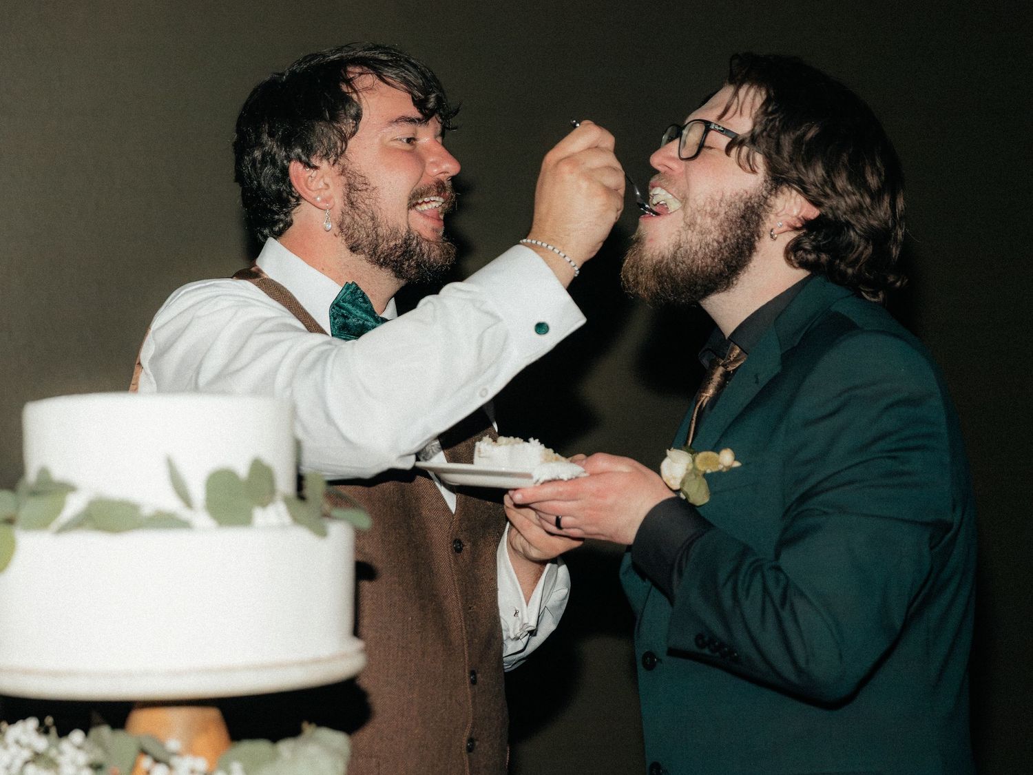 Two grooms share a joyful cake-cutting moment at their wedding reception, feeding each other white cake.