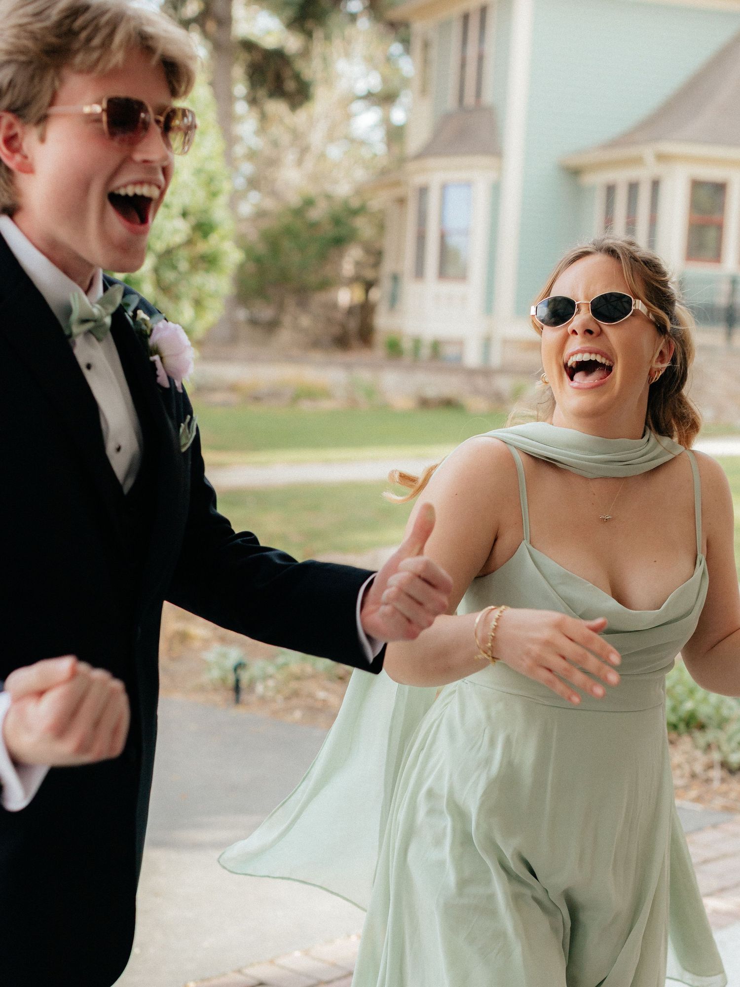 Joyful couple in formal attire laughing outdoors, woman in mint green gown and man in black tuxedo with green tie.