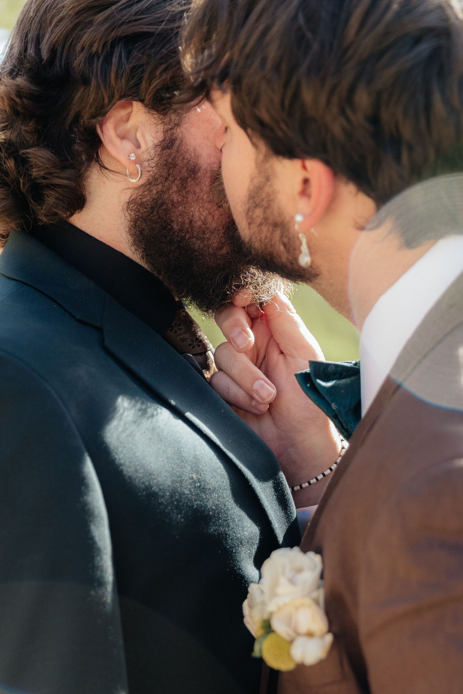 Two grooms share an intimate moment, one kissing the others hand, both wearing suits with boutonnieres at their wedding.