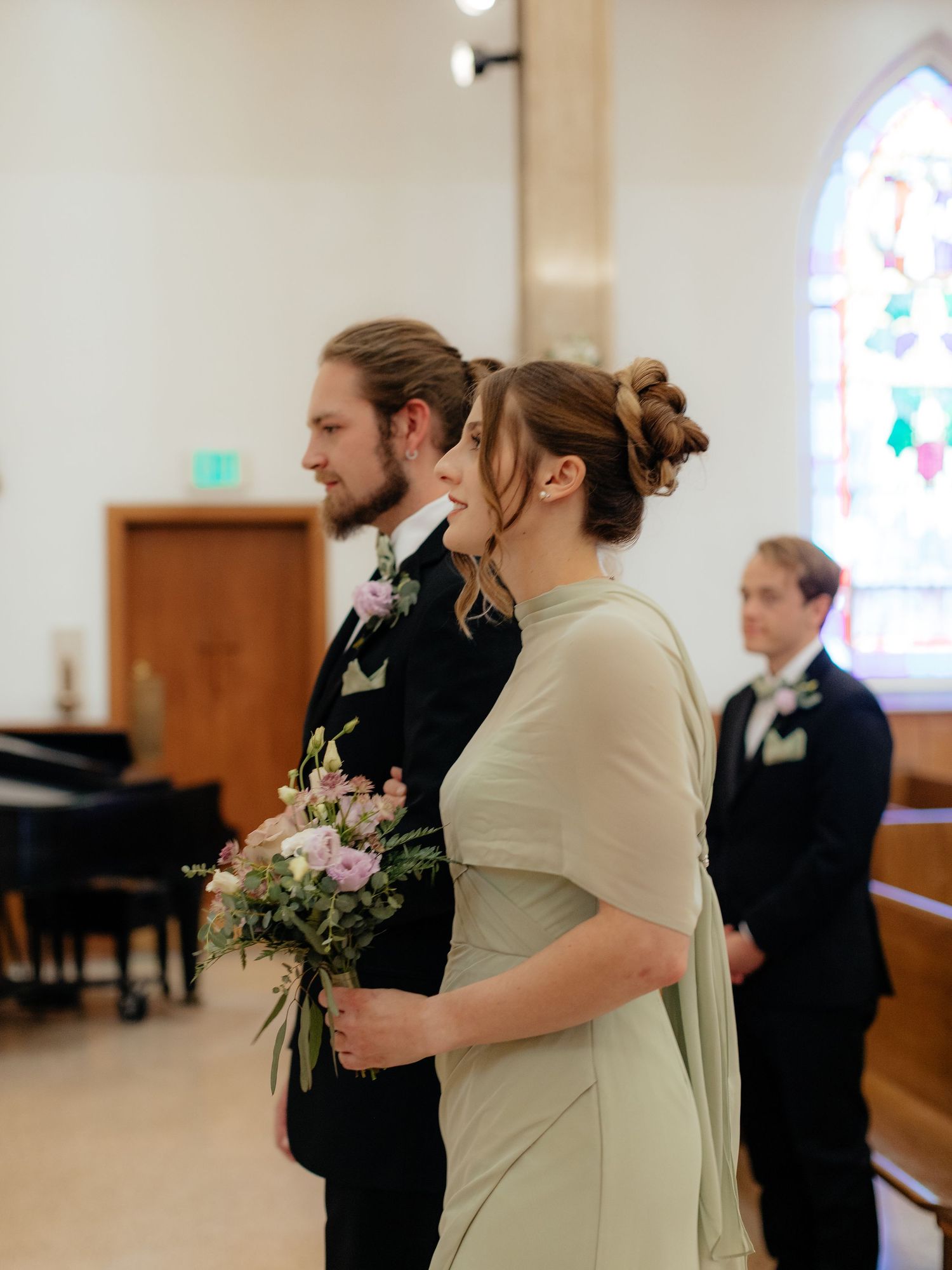 Smiling bride holding pink floral bouquet stands beside groom in black suit inside church ceremony.