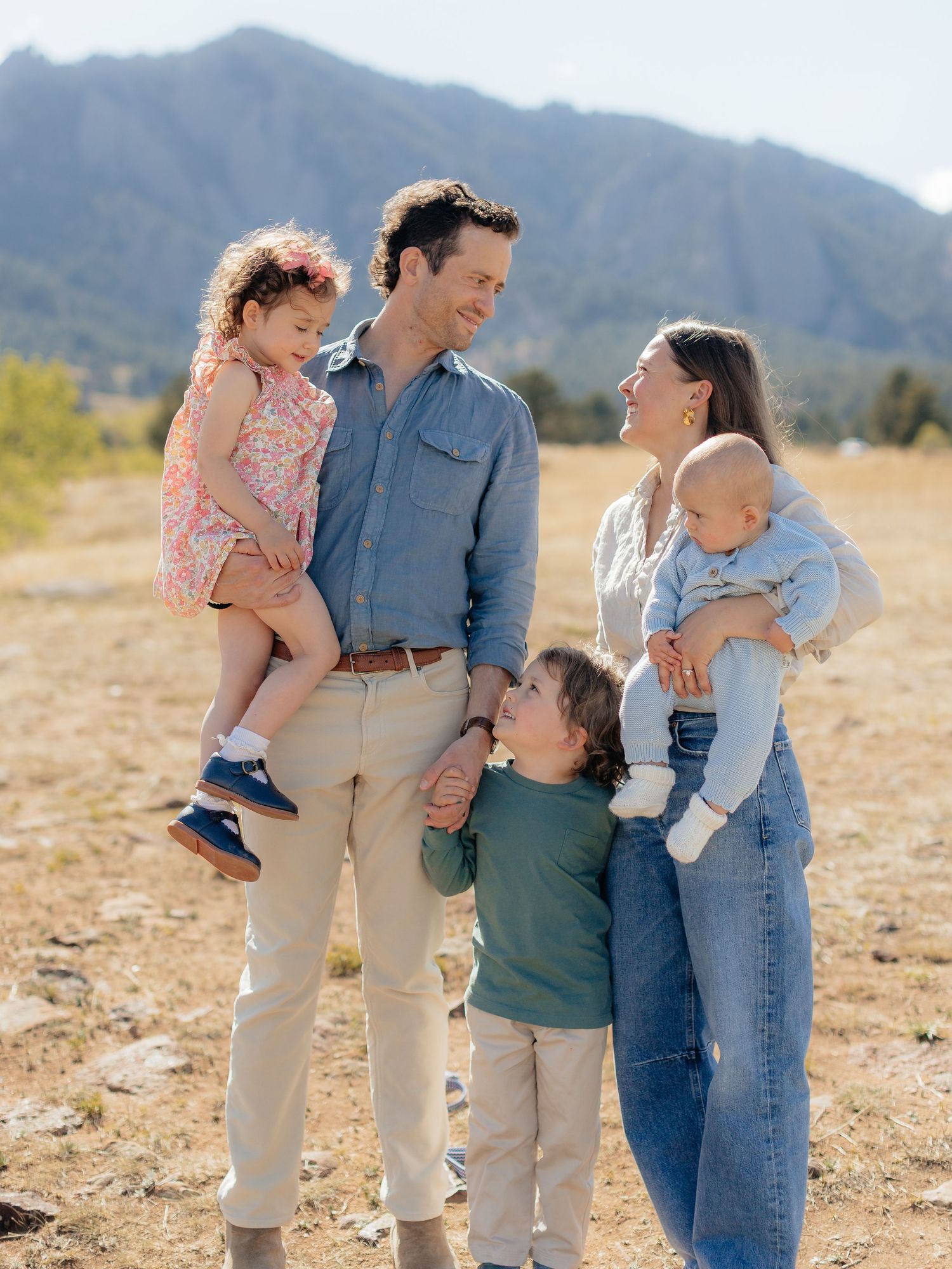 Happy family of five posing outdoors in a sunny field with mountains in the background.
