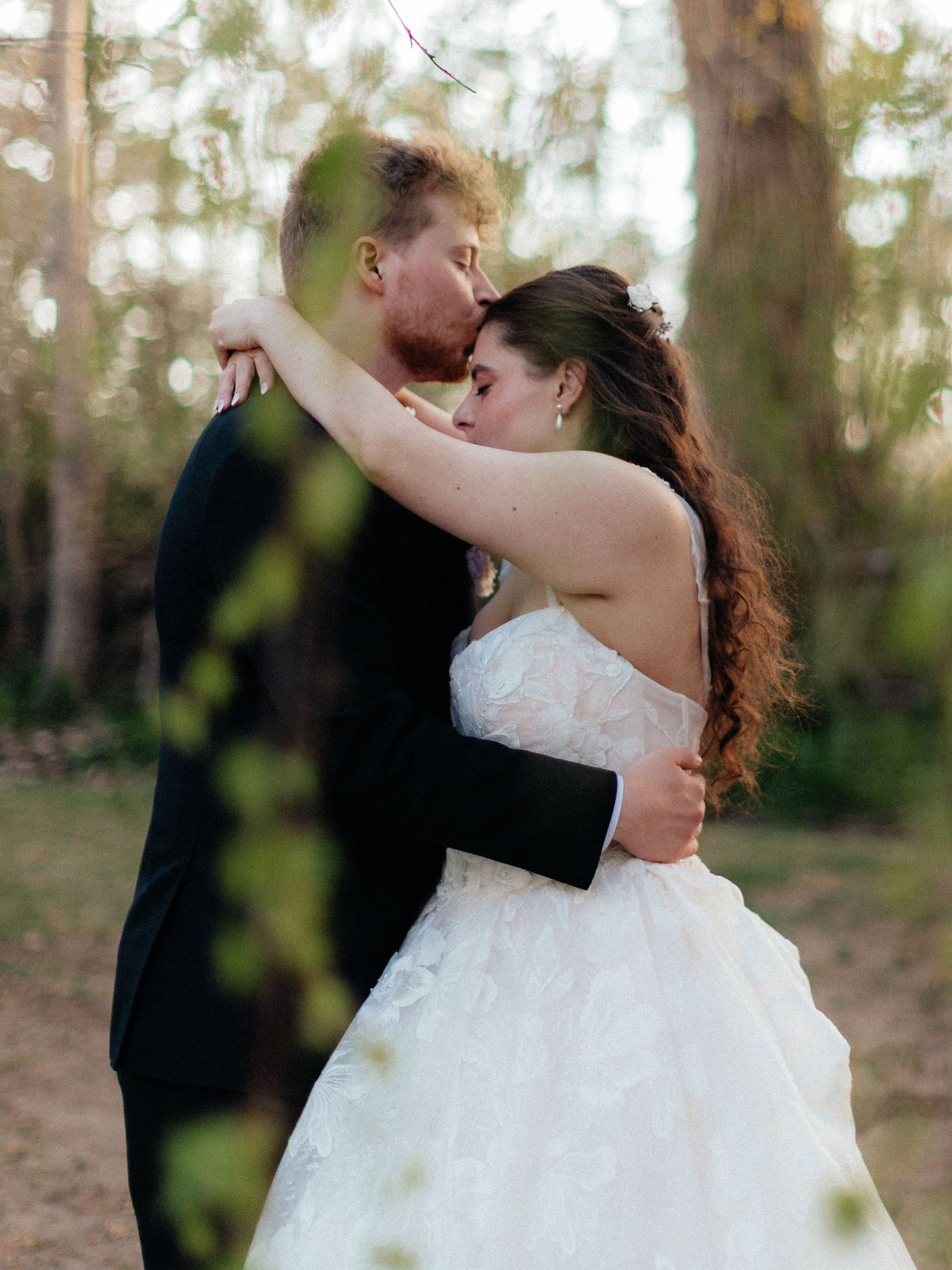 Bride and groom share a romantic kiss outdoors surrounded by trees in a lush forest wedding setting.