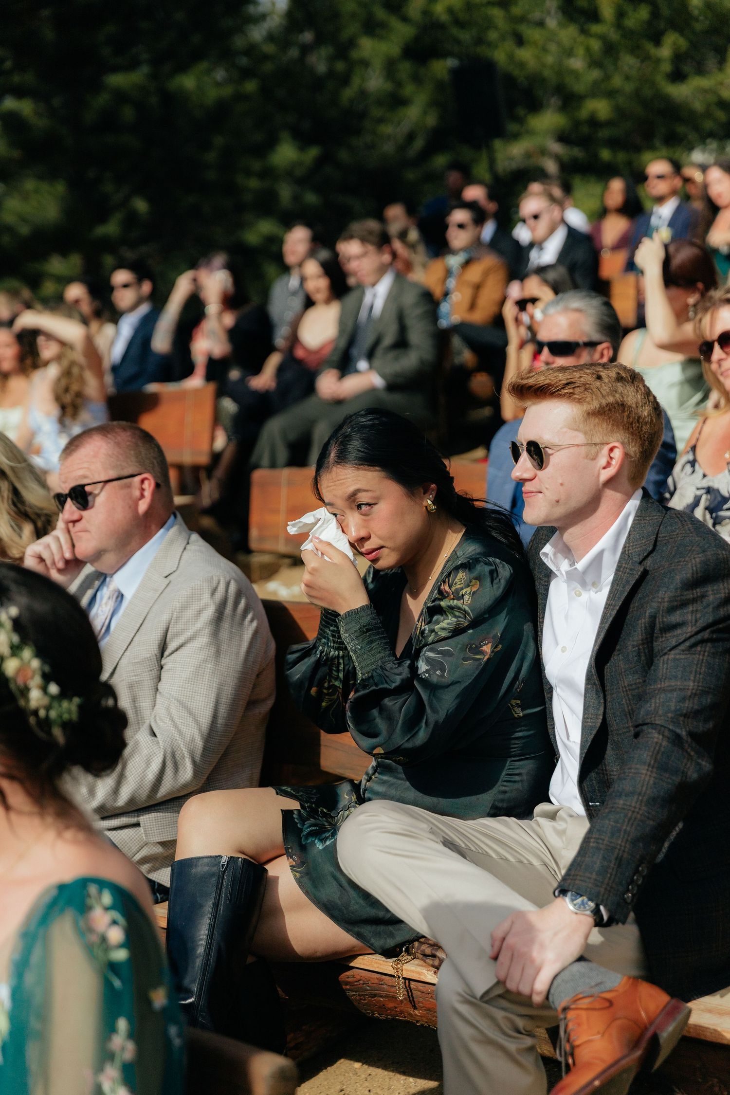Emotional wedding guest wiping tears during outdoor ceremony seated on wooden benches in sunlight.