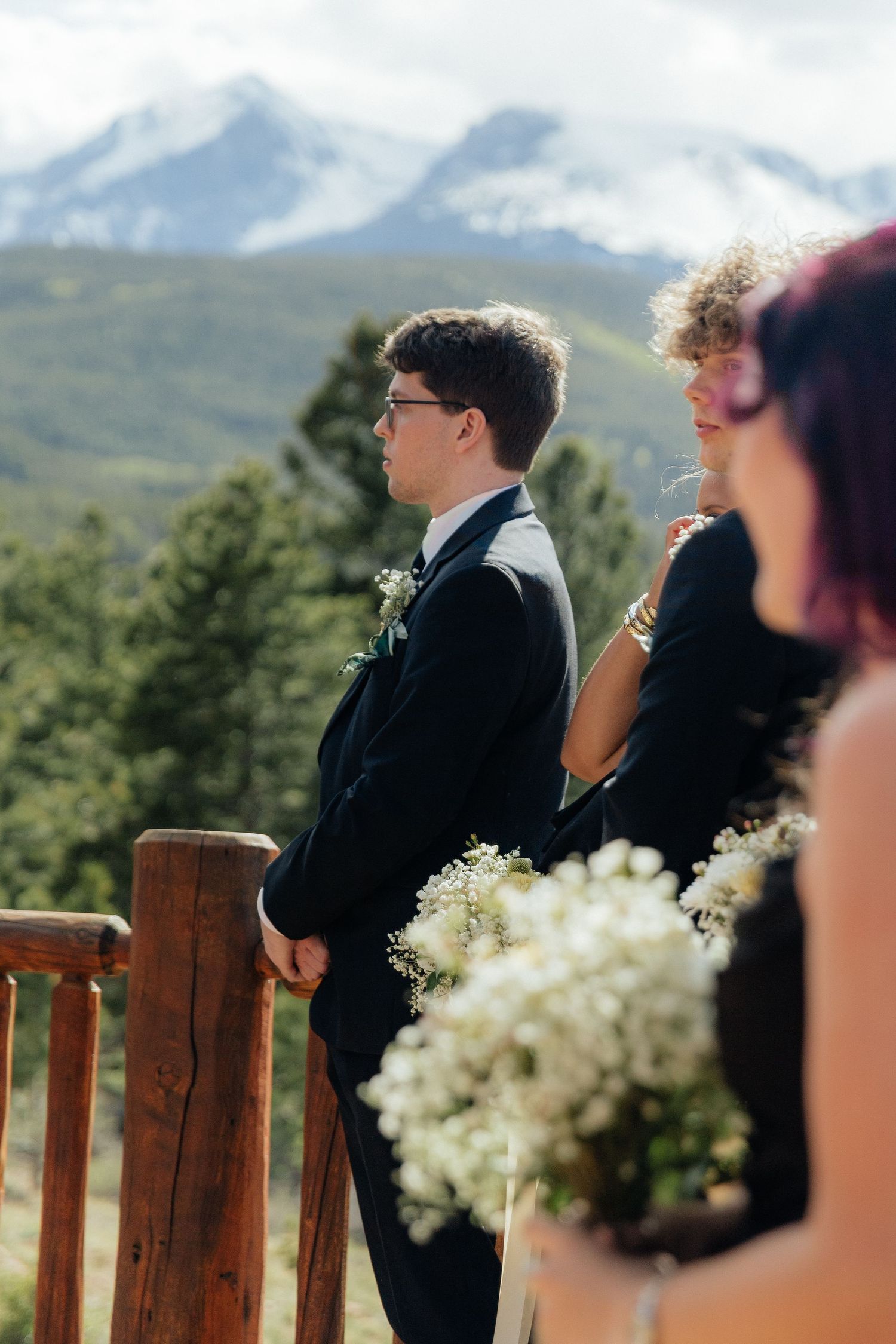 Groom in black suit stands at wooden railing with mountain backdrop during outdoor wedding ceremony.