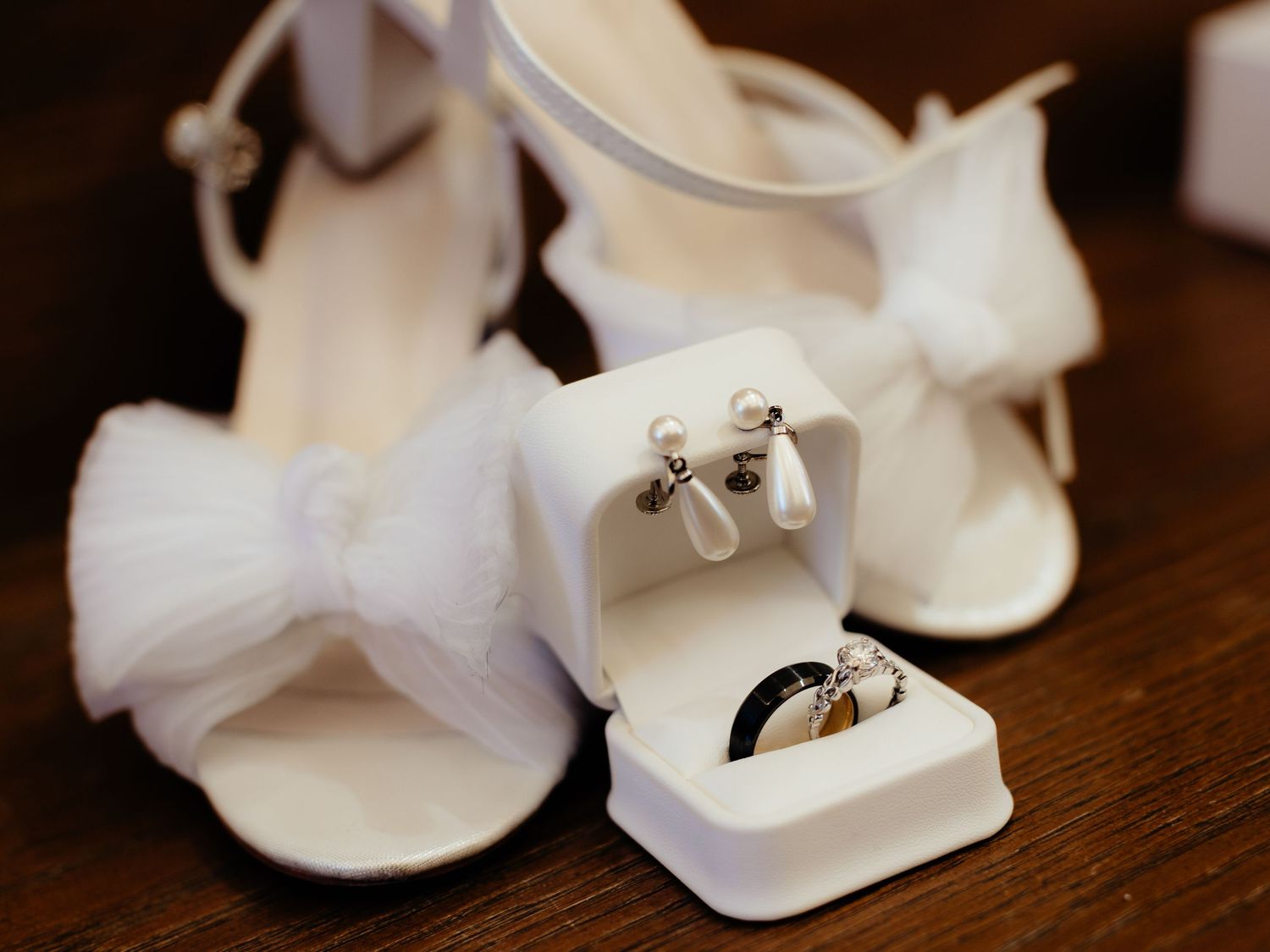 White bridal heels with bow detail beside an open jewelry box with pearl earrings and wedding rings on wooden surface.