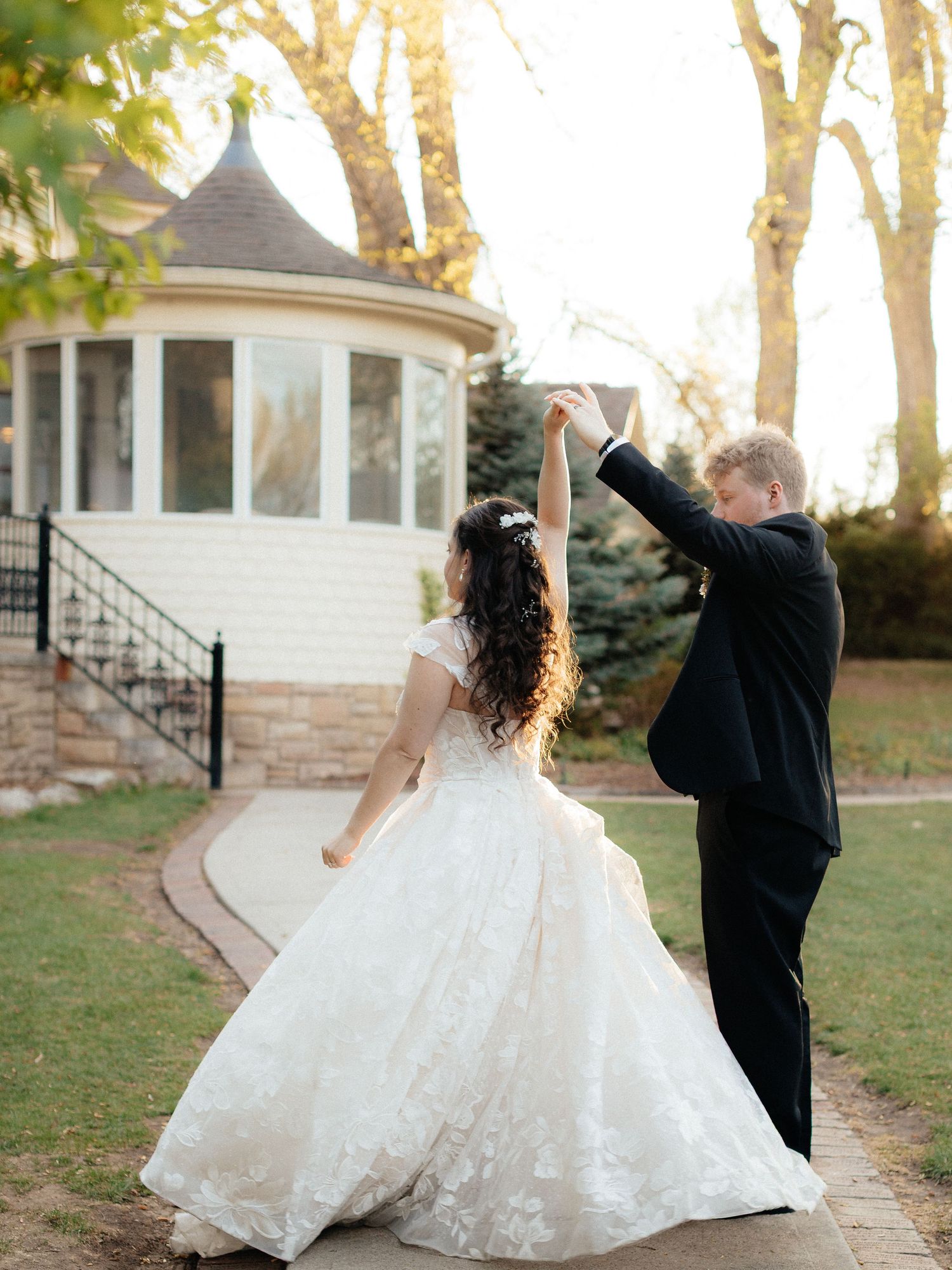 Bride in lace ball gown twirls as groom in black tuxedo lifts her hand outside a white gazebo.
