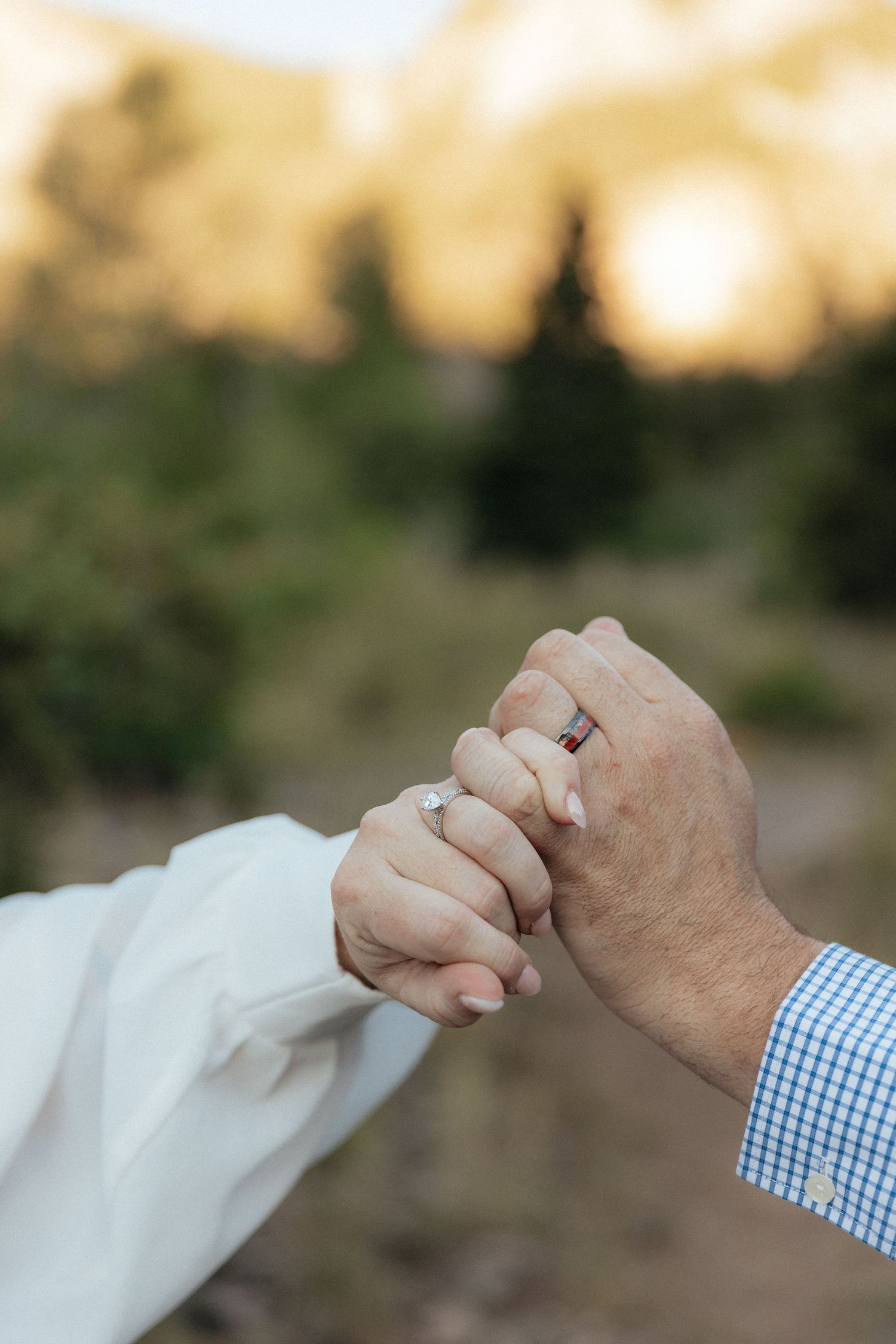 Close-up of newlyweds holding hands with wedding rings, set against a blurred mountain and forest backdrop at sunset.