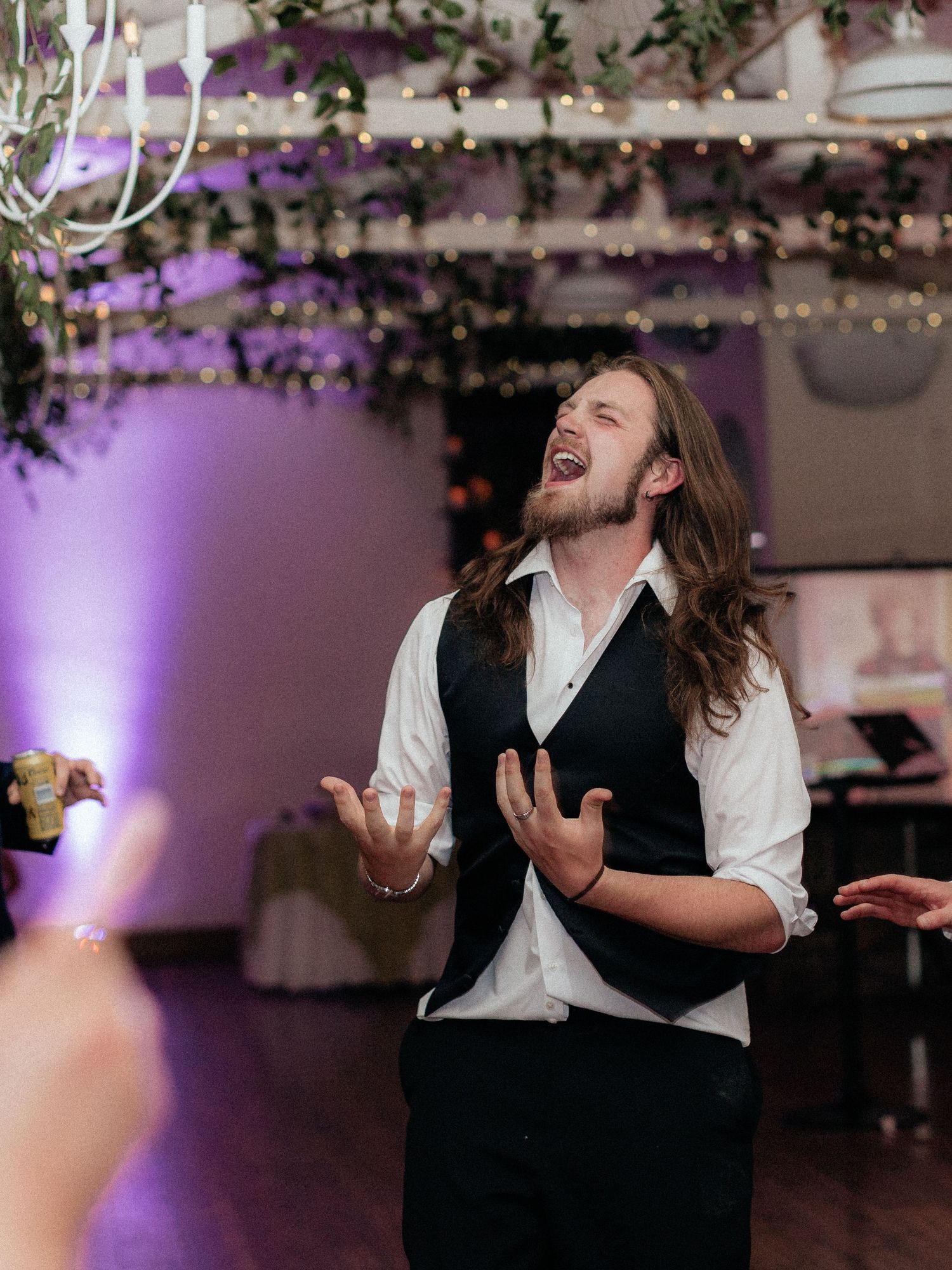 Long-haired groom singing joyfully at wedding reception with purple uplighting and string lights.