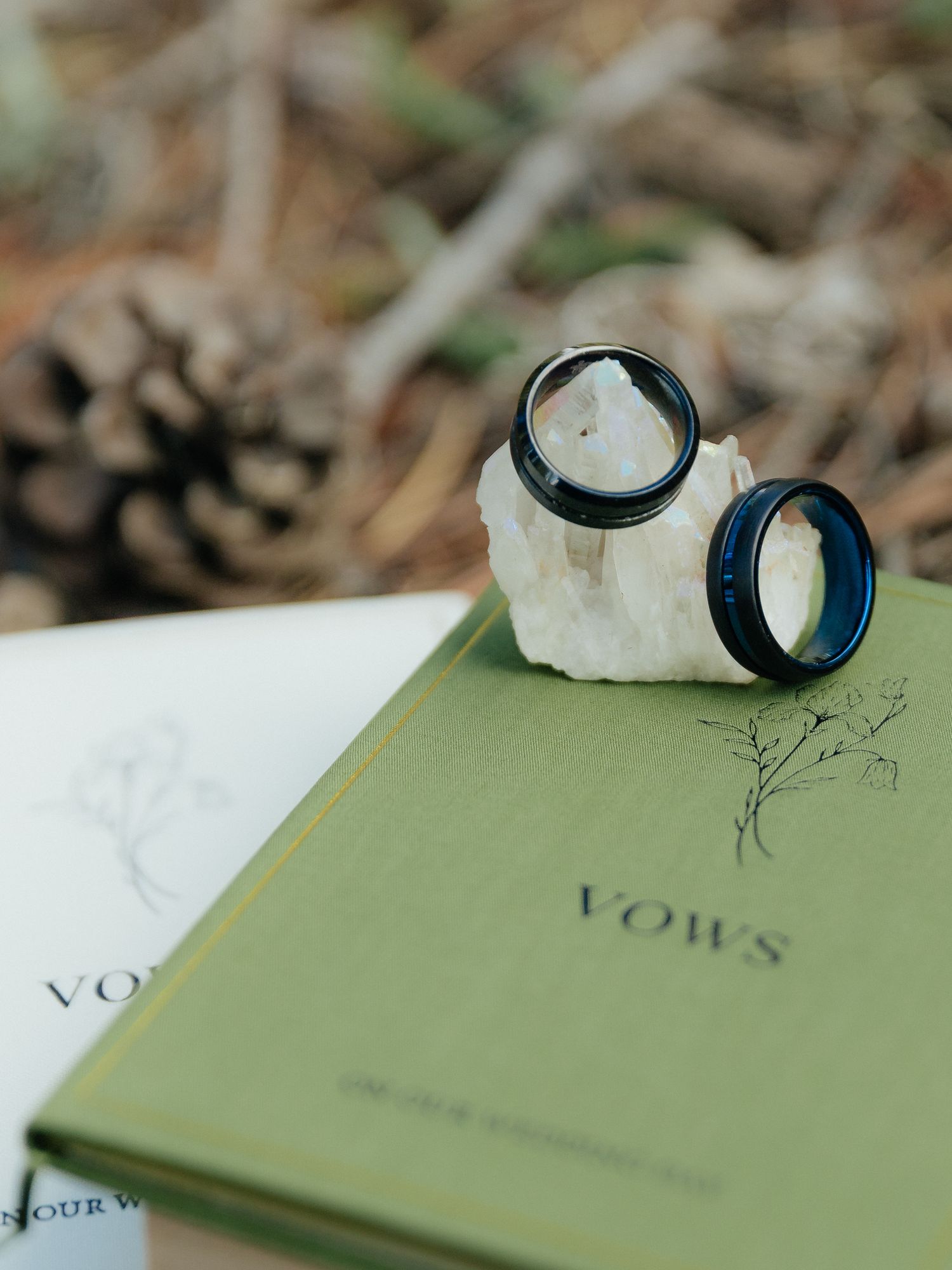 Two black wedding bands rest on a white stone atop a green vows book outdoors.