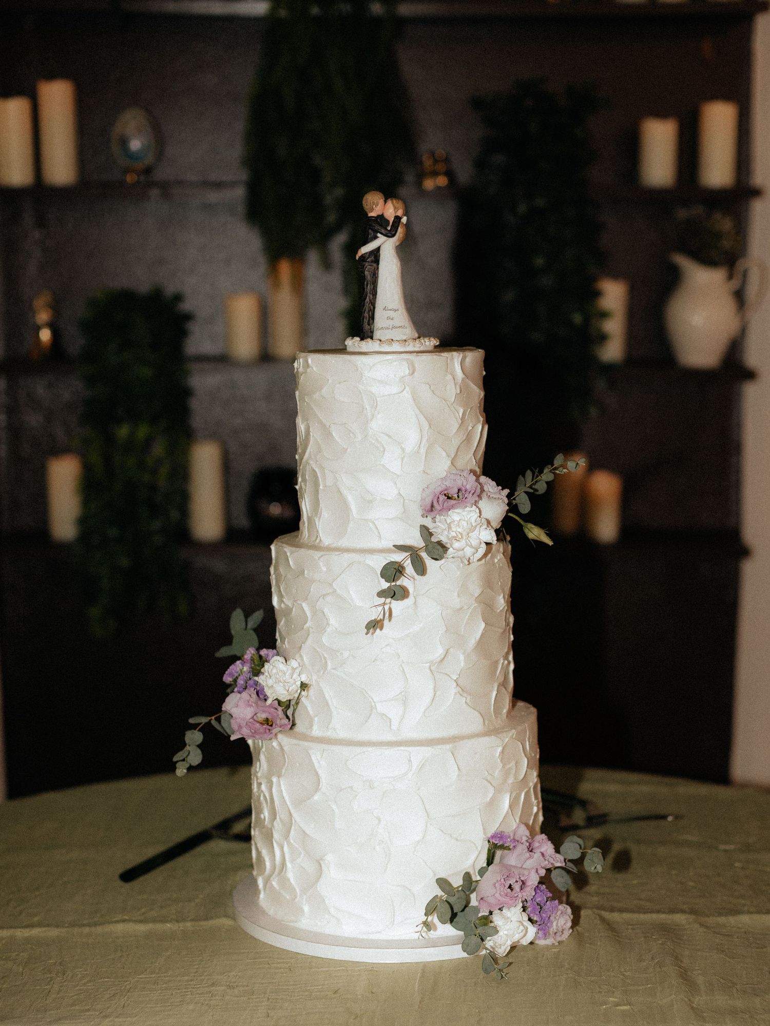 Four-tier white textured wedding cake with purple floral accents and a bride and groom cake topper on a draped table.