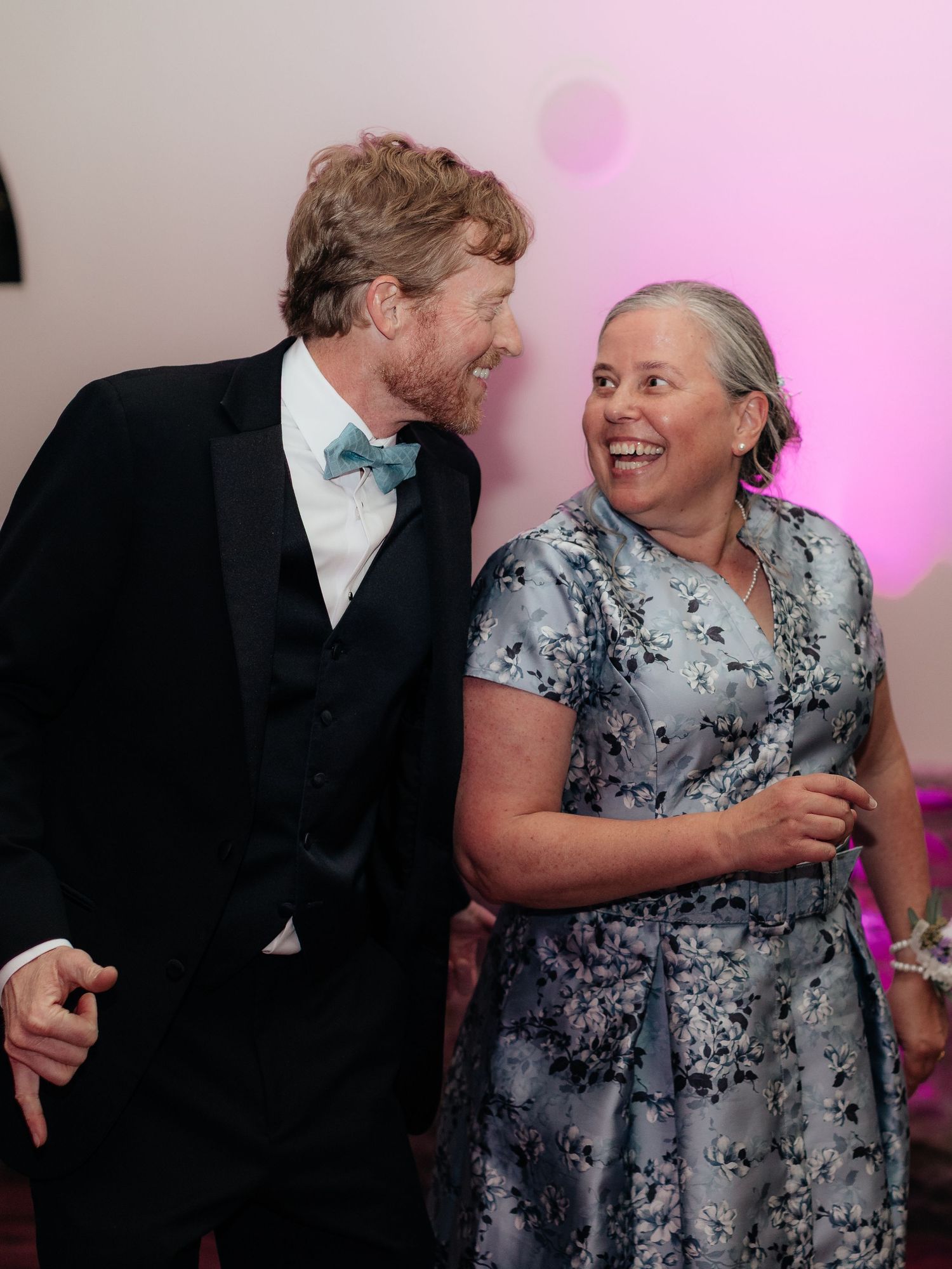 A man in a tuxedo with bow tie and a woman in floral dress laughing together at a formal event.