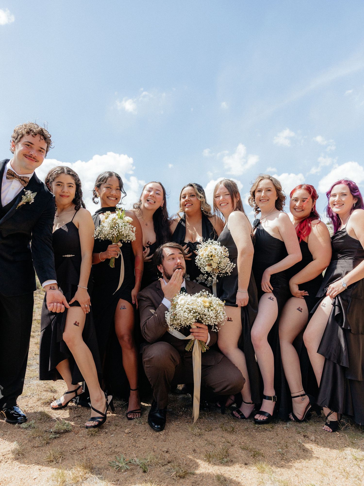 Wedding party in black formal attire poses outdoors, holding white bouquets under a bright blue sky.