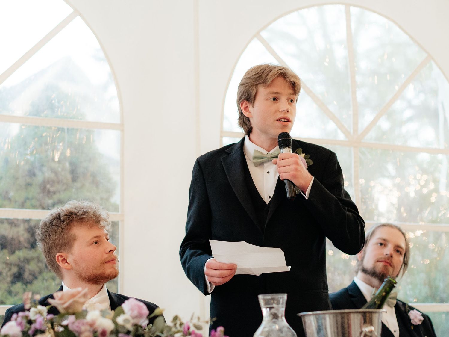 Young man in tuxedo giving wedding speech at reception, holding paper and microphone near floral arrangements.
