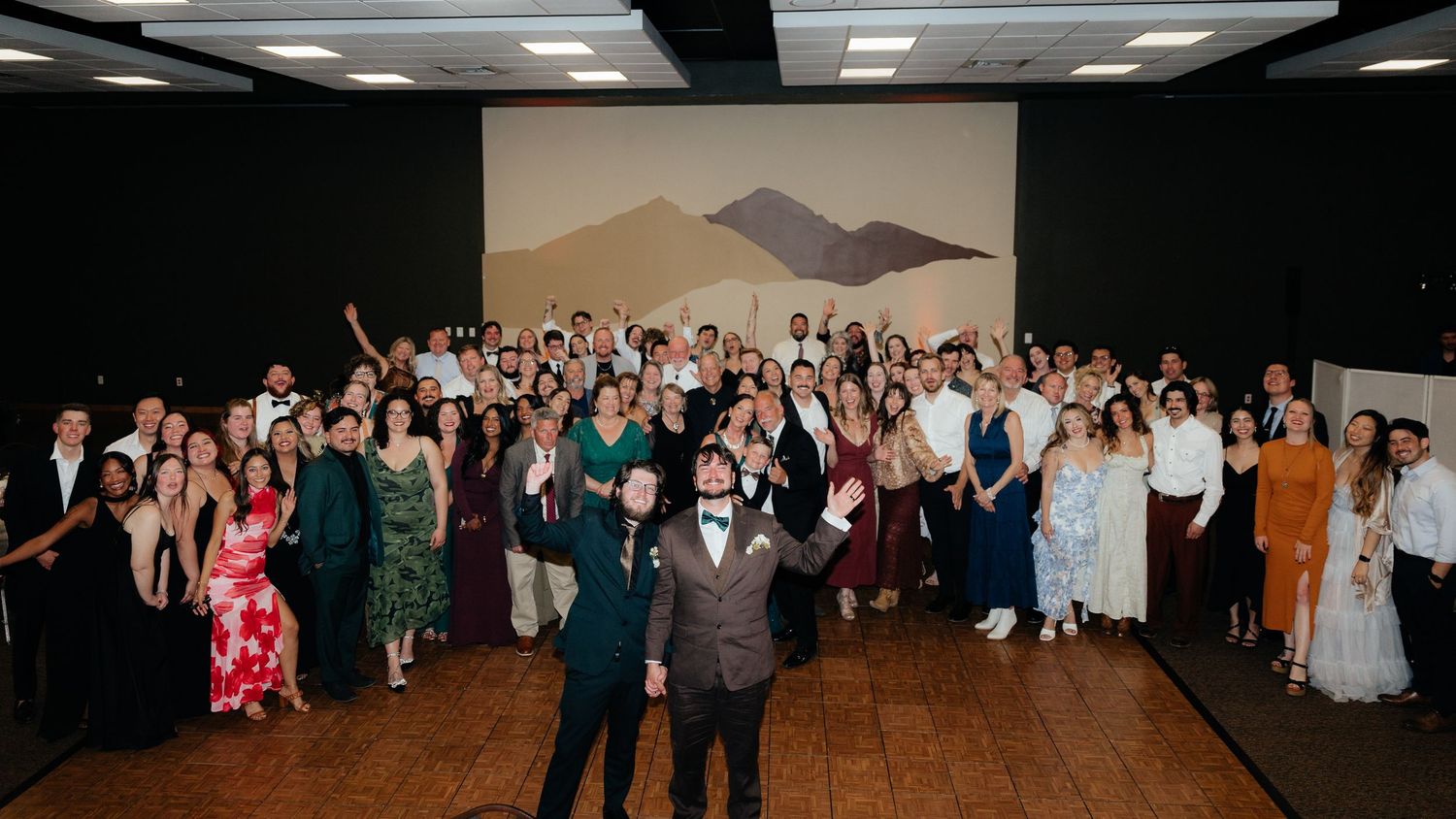 Large wedding reception group photo with guests in formal attire posing together in a decorated event hall.
