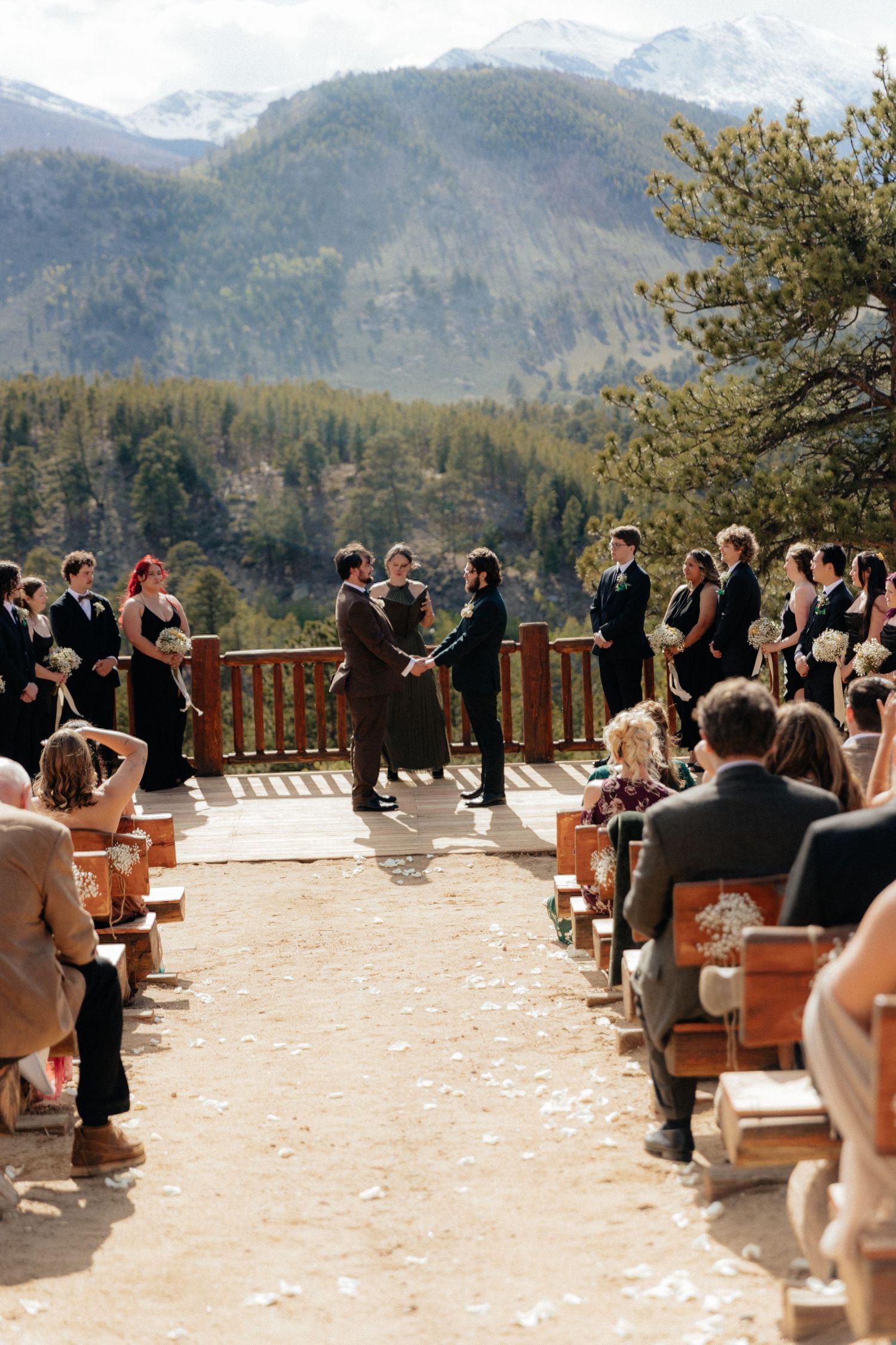 Outdoor mountain wedding ceremony on a rustic deck with guests seated along the aisle, pine trees and snowy peaks in background.