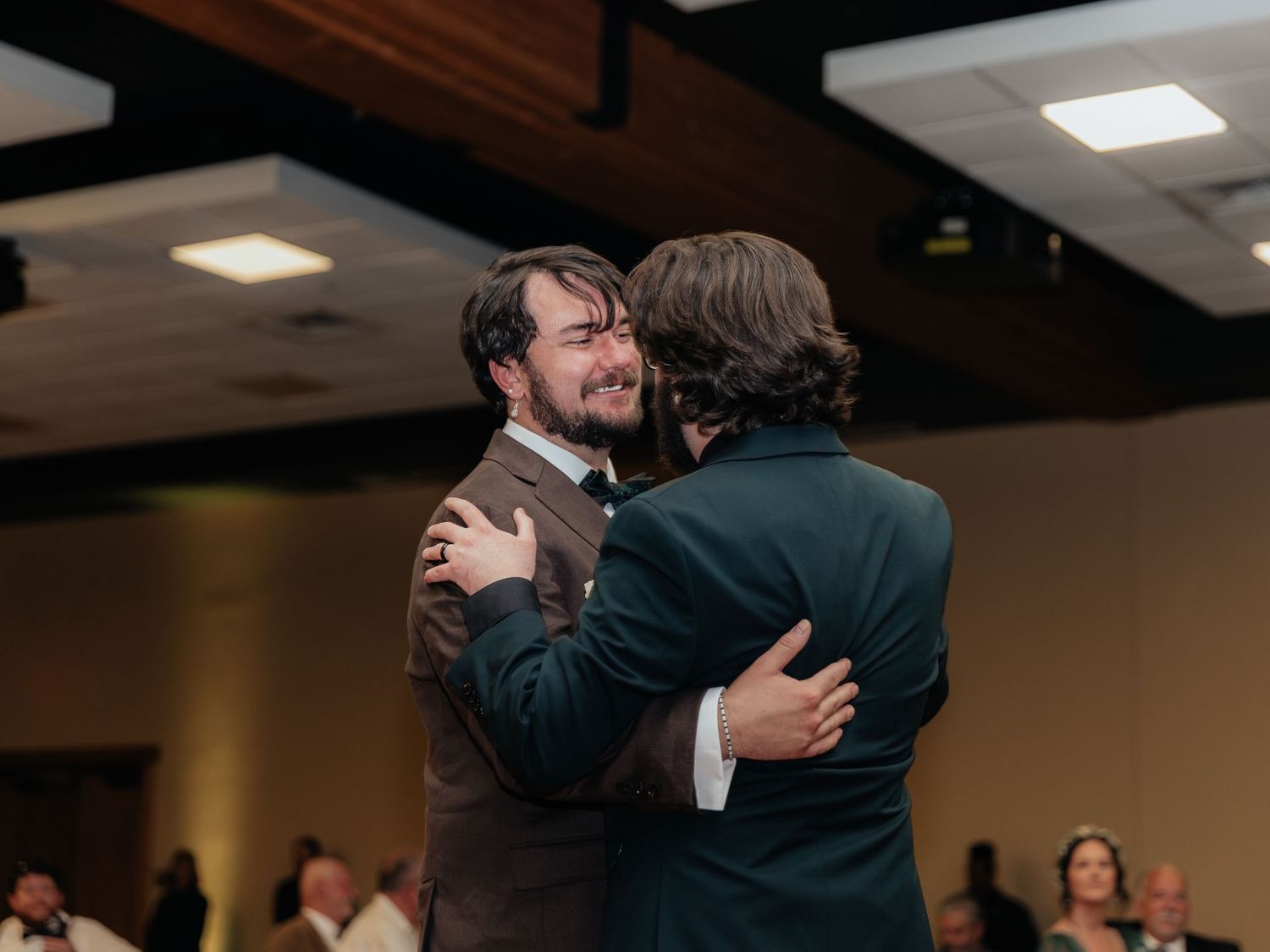 Two men share an emotional hug on a wedding dance floor, dressed in brown and teal suits.