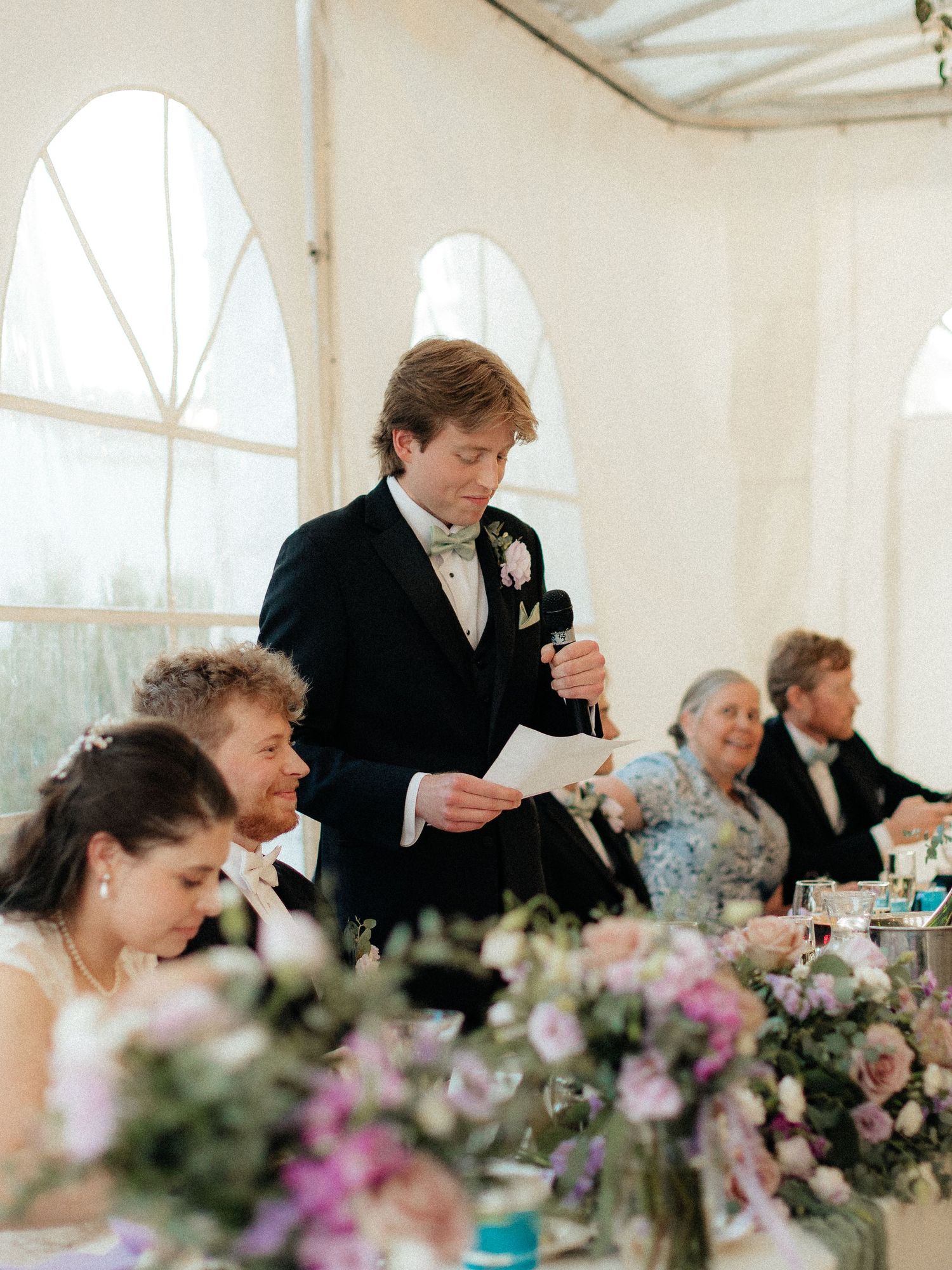 Groom delivering wedding speech at elegant marquee reception with floral centerpieces and seated guests.