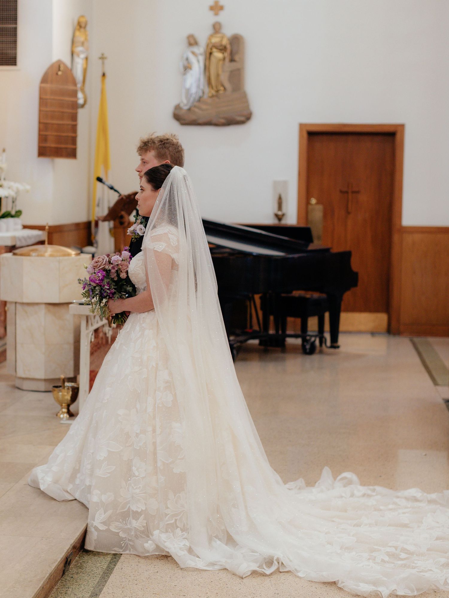 Bride in elegant lace gown with long cathedral veil stands at church altar holding purple bouquet.