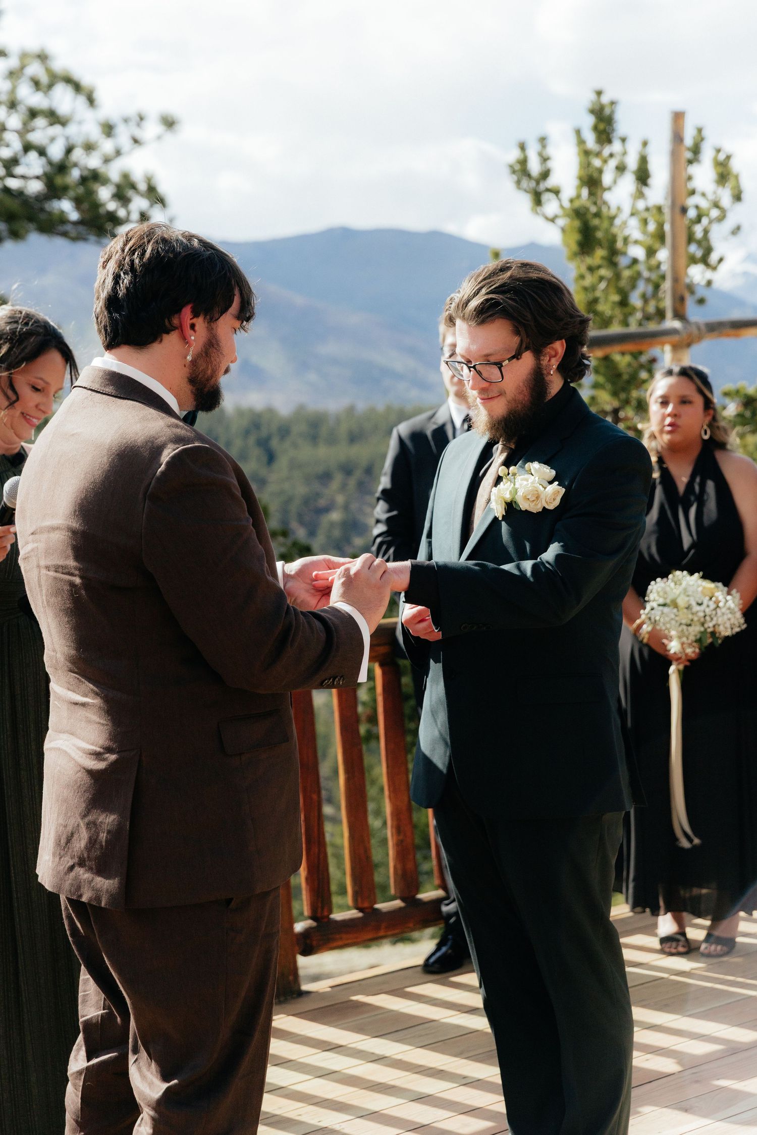 Two grooms exchanging rings during wedding ceremony on mountain deck with bridal party behind.